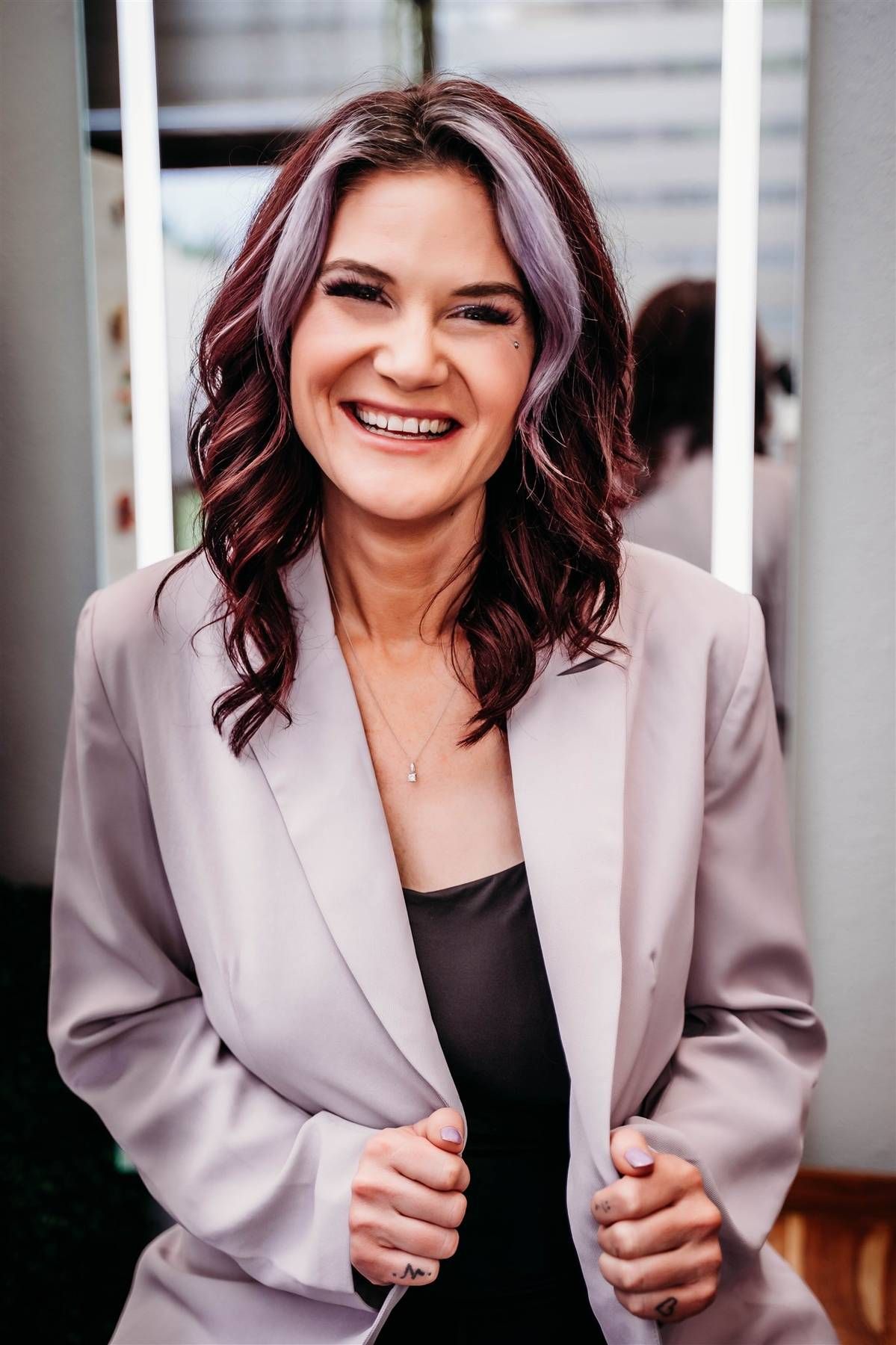Happy woman with purple-highlighted dark hair laughing, wearing a light grey blazer over a black top.