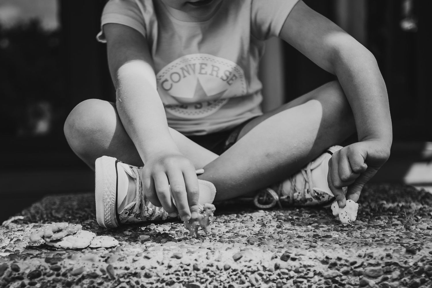 Black and white photo of a child in Converse sneakers sitting cross-legged on a textured surface.