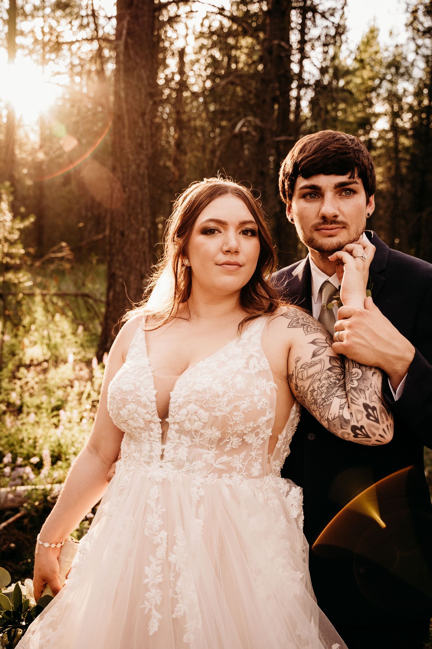 Bride in deep V lace gown and tattooed groom pose in sun-drenched forest during wedding portraits.