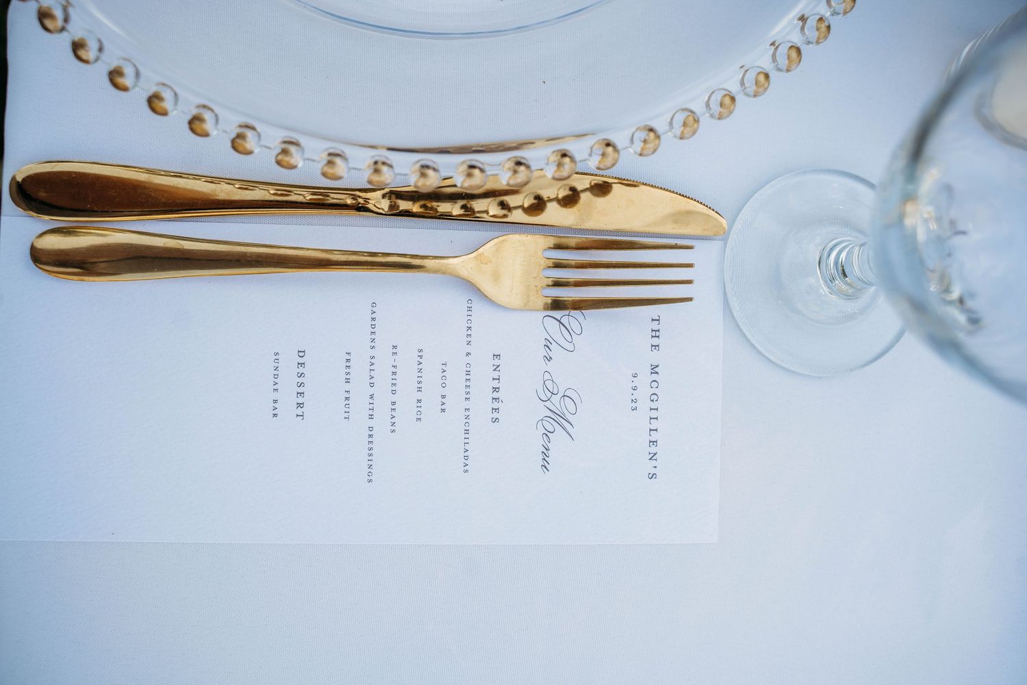 Gold fork and knife beside a printed wedding menu card on a white tablecloth with beaded charger plate.