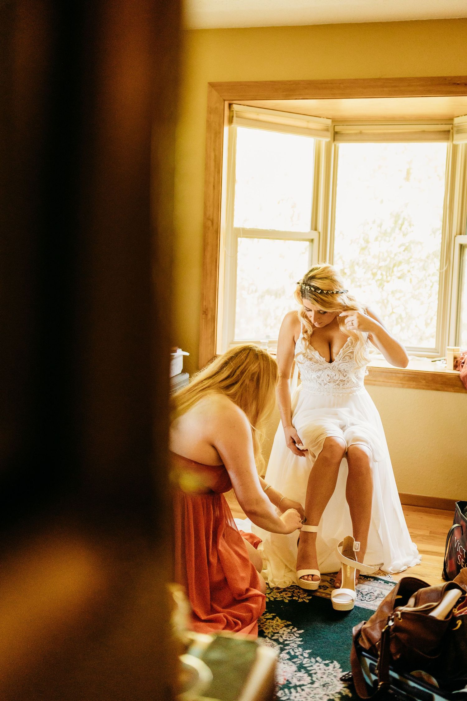 Bridesmaid helps bride with shoes by sunny window before wedding ceremony.