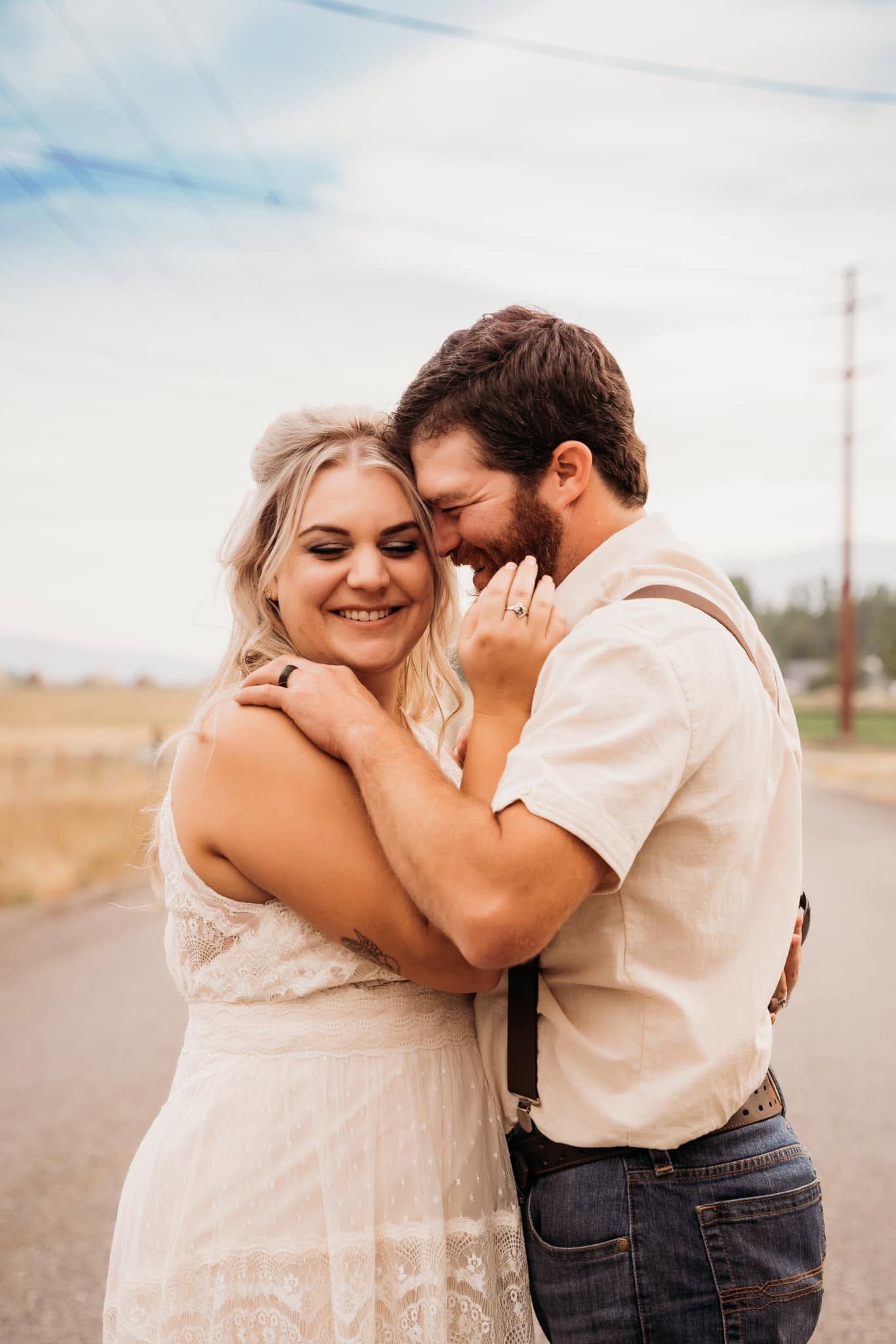 Happy bride and groom embracing on rural road, bride showing off wedding ring in lace dress.