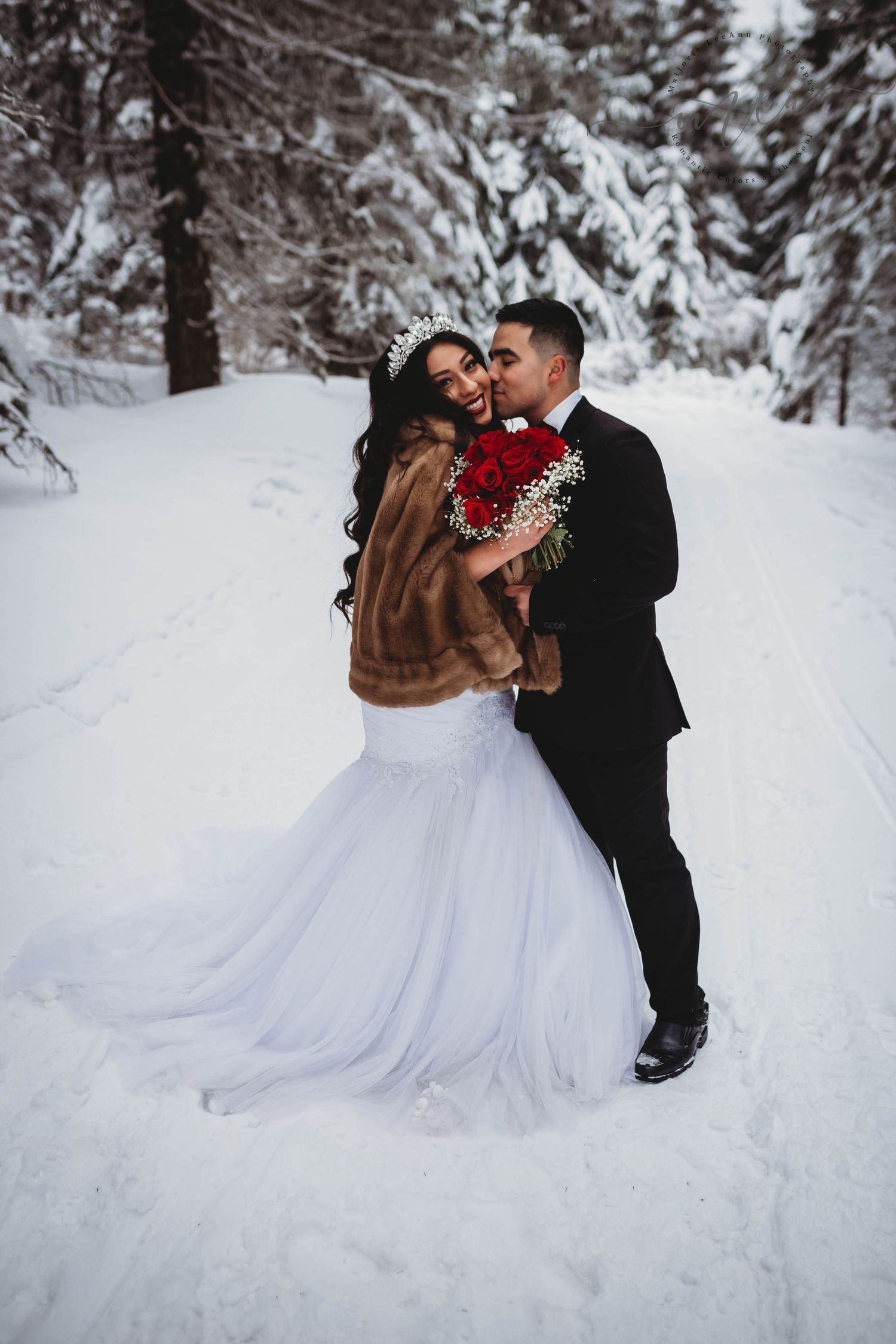 Bride and groom embrace in snowy winter forest with red rose bouquet.