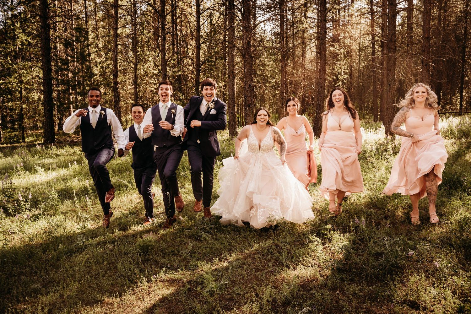 Joyful wedding party running through sunlit forest with bride in ruffled gown and bridesmaids in blush pink dresses.