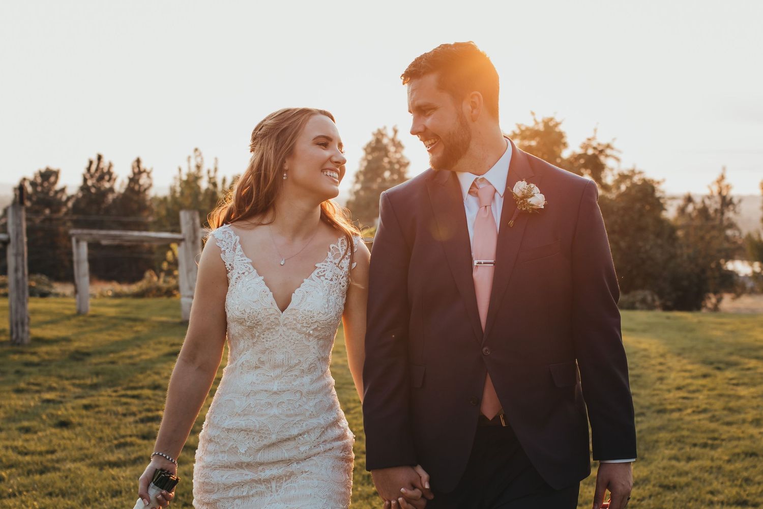 Bride and groom walk hand-in-hand through a sunlit field at golden hour on their wedding day.