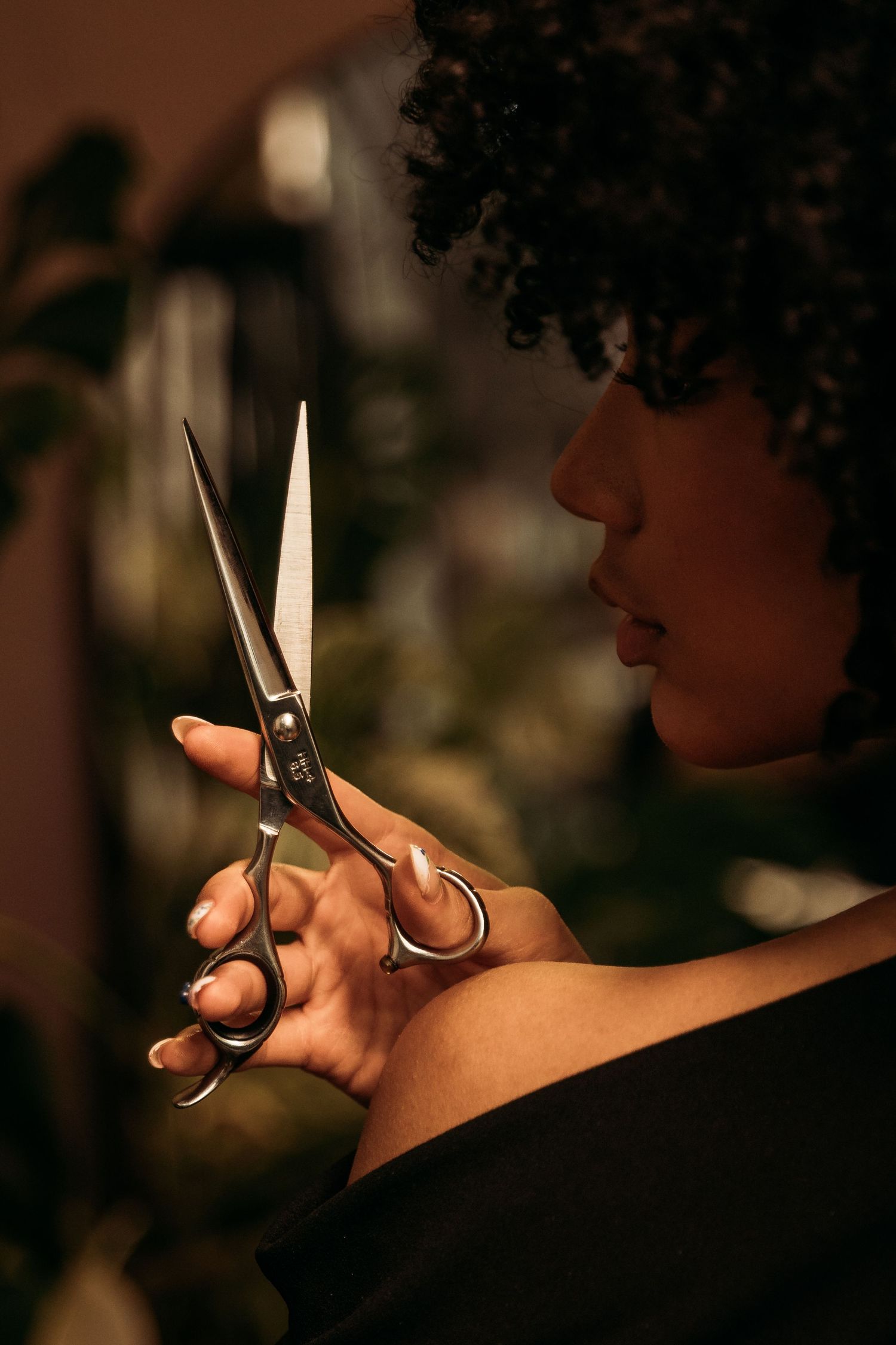 Close-up of a curly-haired woman examining professional hairdressing scissors in dim warm lighting.