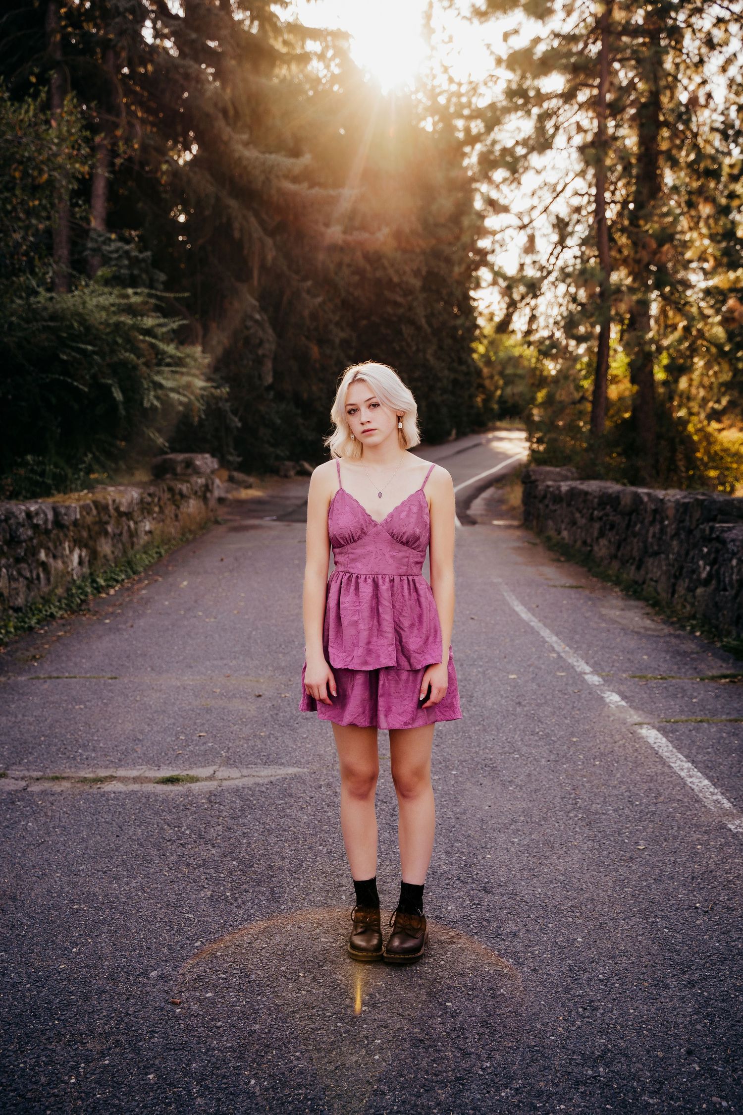 Young blonde woman in pink sundress and boots standing on a tree-lined road at golden hour.