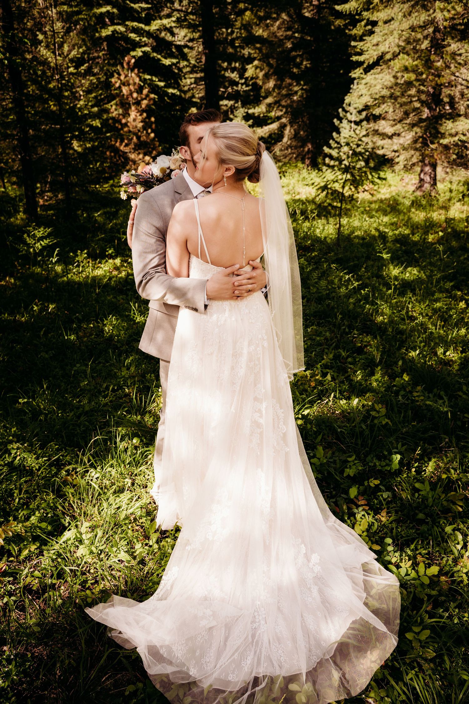 Bride and groom sharing a kiss in a sunlit forest, bride wearing a backless lace wedding gown.