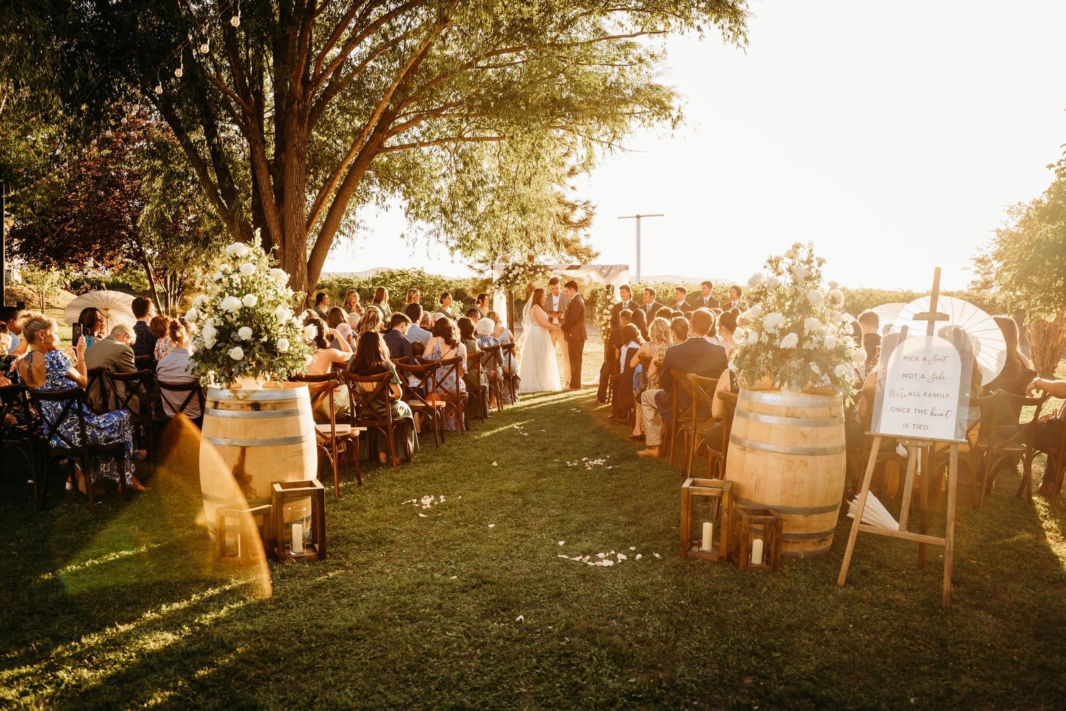 Outdoor wedding ceremony at golden hour with guests seated along a flower-lined aisle near wine barrels.
