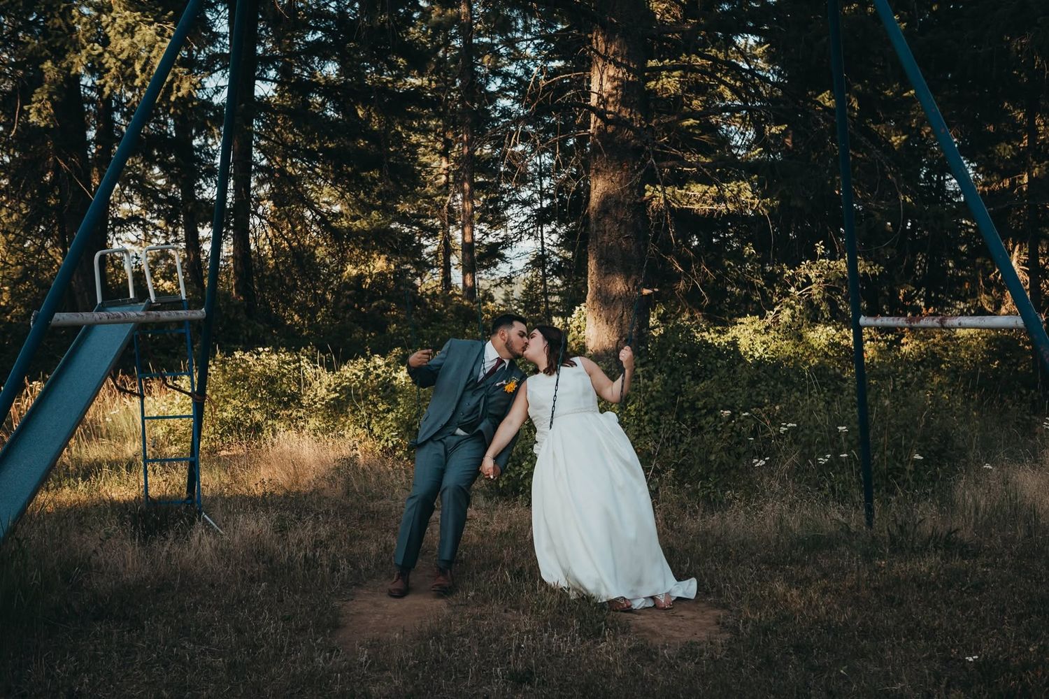 Bride and groom sharing a romantic kiss near a swing set in a forest setting.