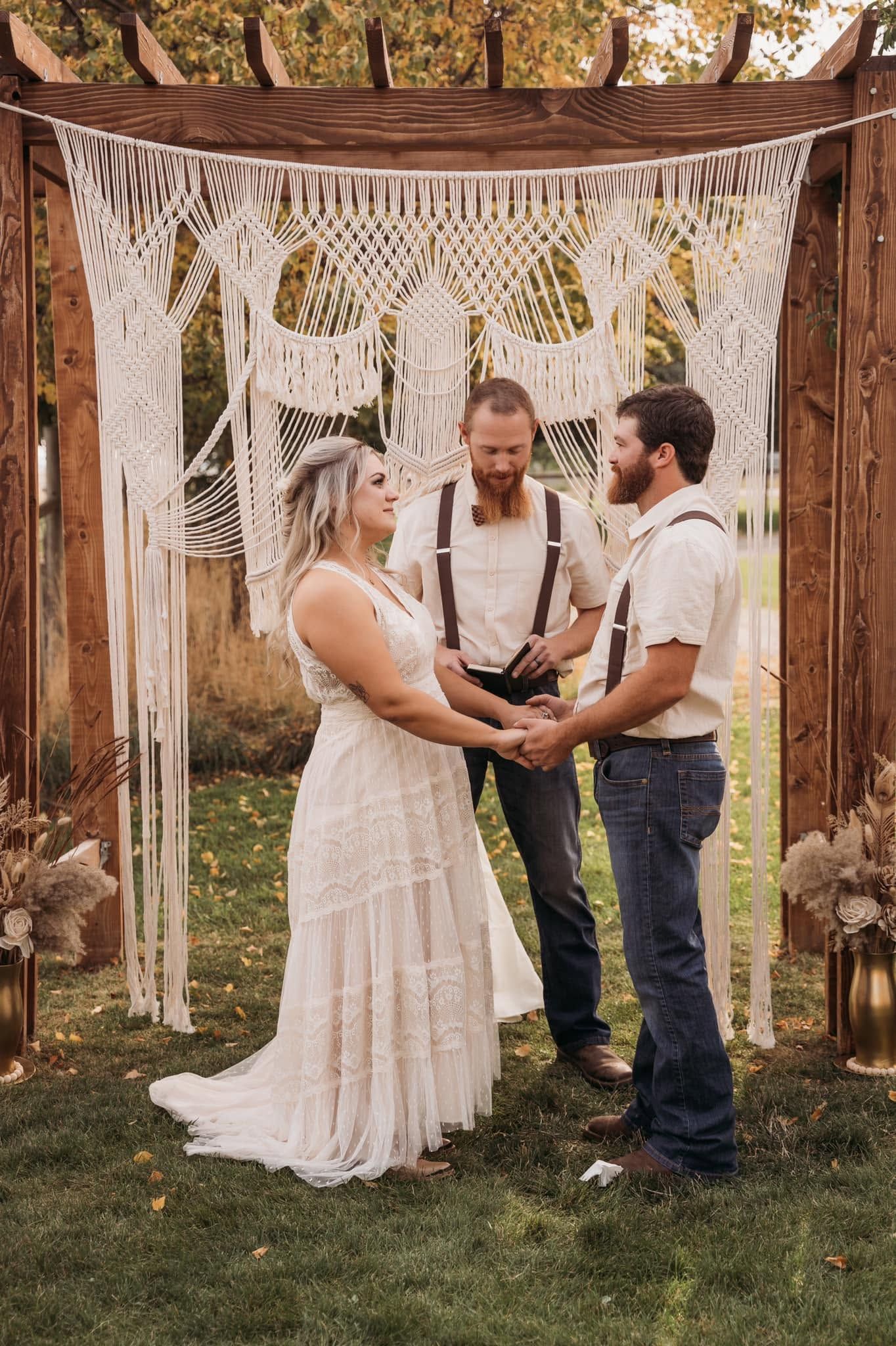 Bride and groom exchanging vows under macrame arch with officiant at outdoor boho wedding.