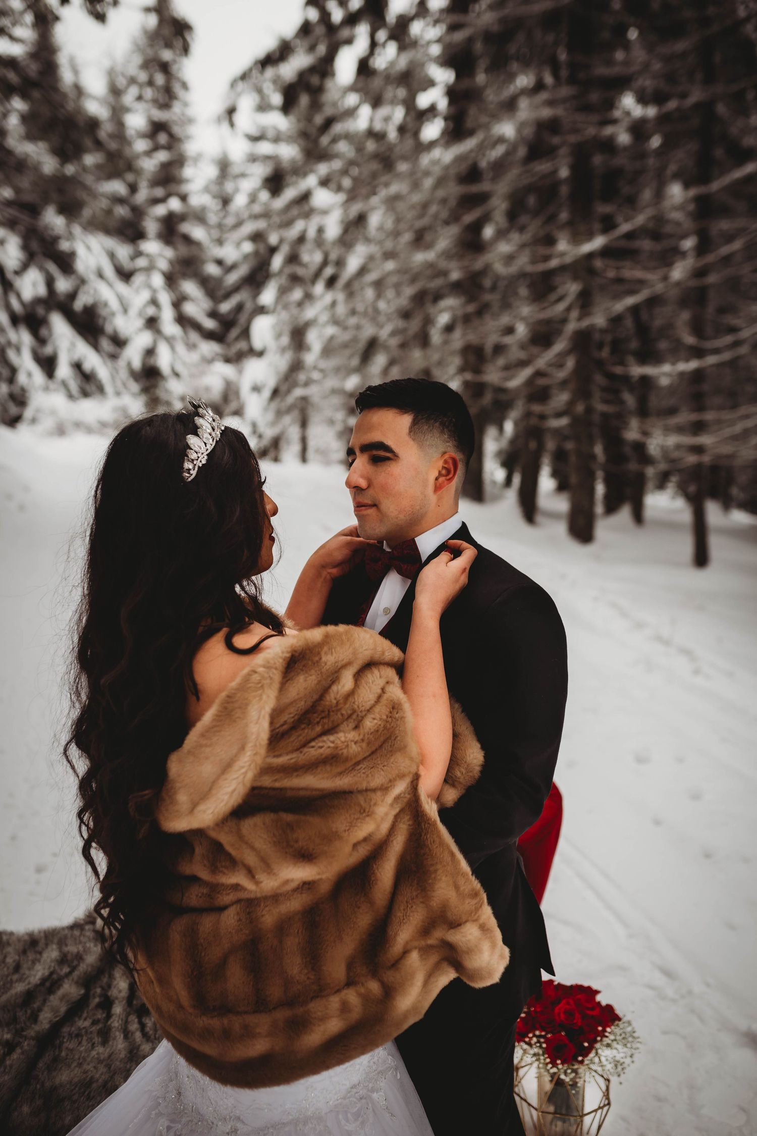 Bride in fur stole adjusts grooms bow tie while standing together on snowy forest path in winter.