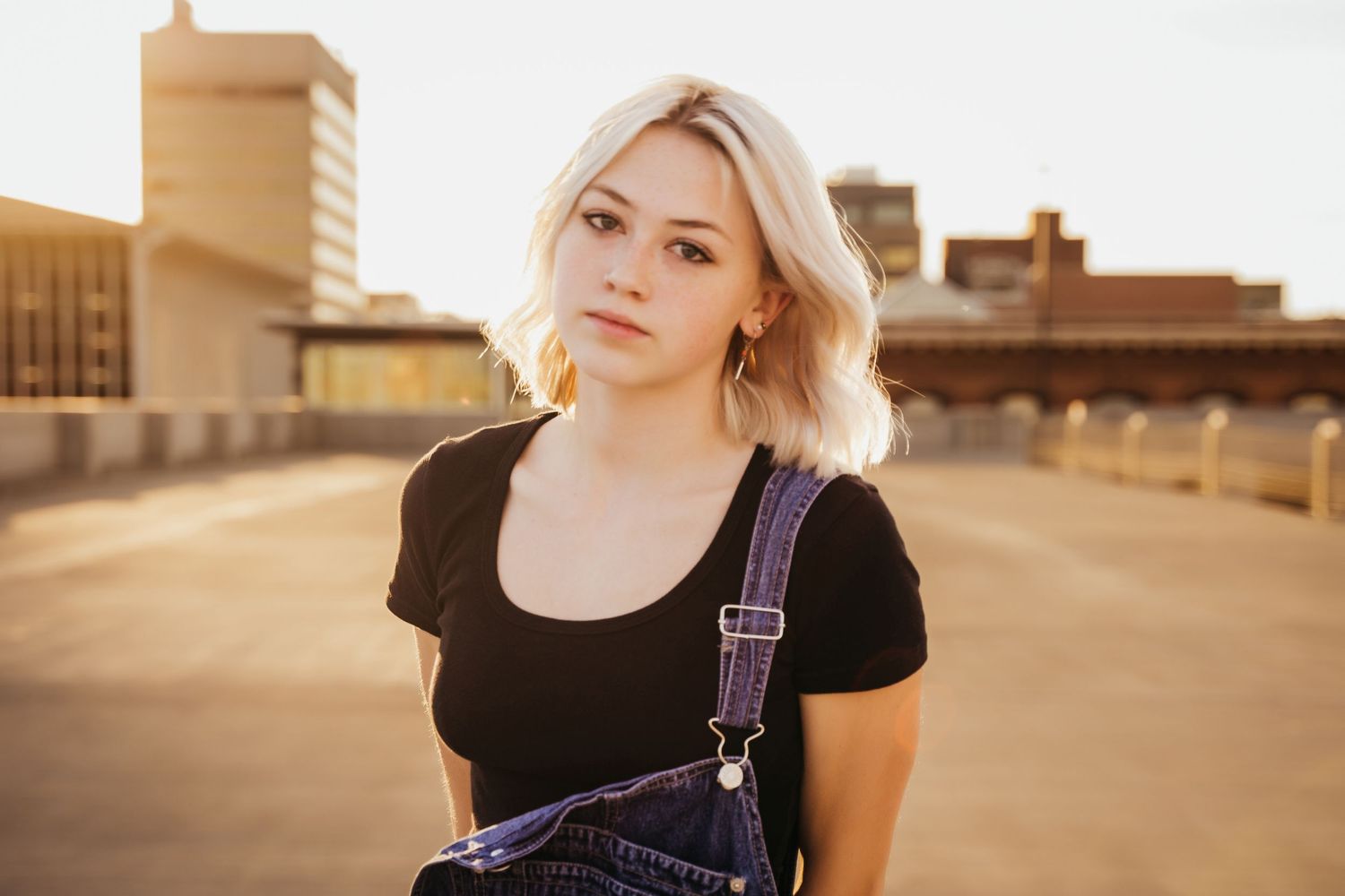 Close-up portrait of blonde woman in black tee and denim overalls on rooftop at golden hour.