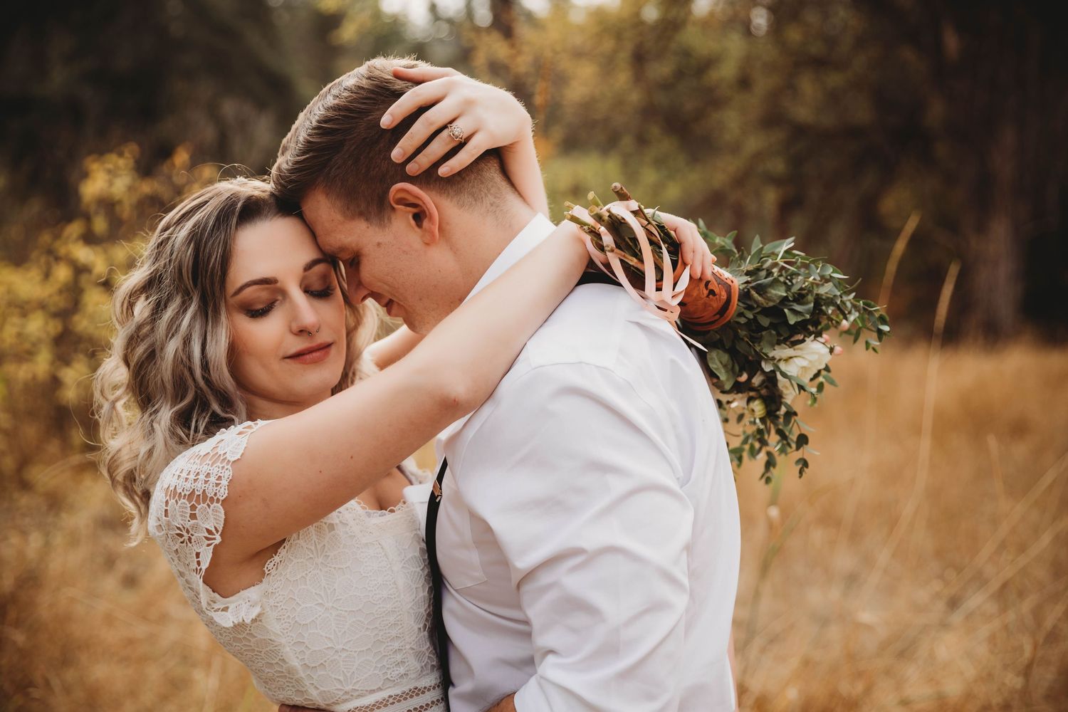 Bride and groom embracing in golden field, groom holding greenery bouquet in autumn forest.