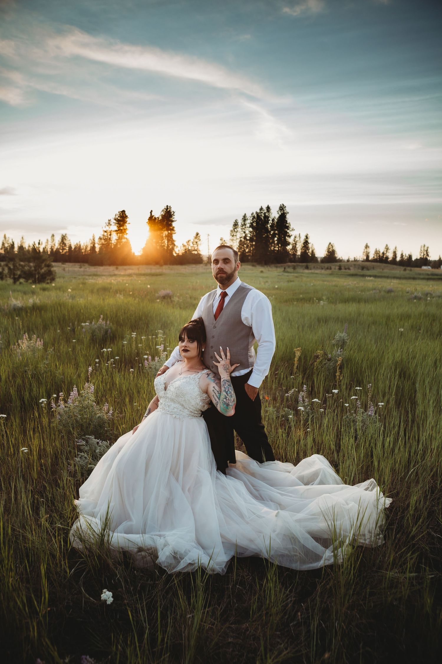 Bride and groom pose in a sunlit meadow at golden hour, her white gown spread across the wildflowers.