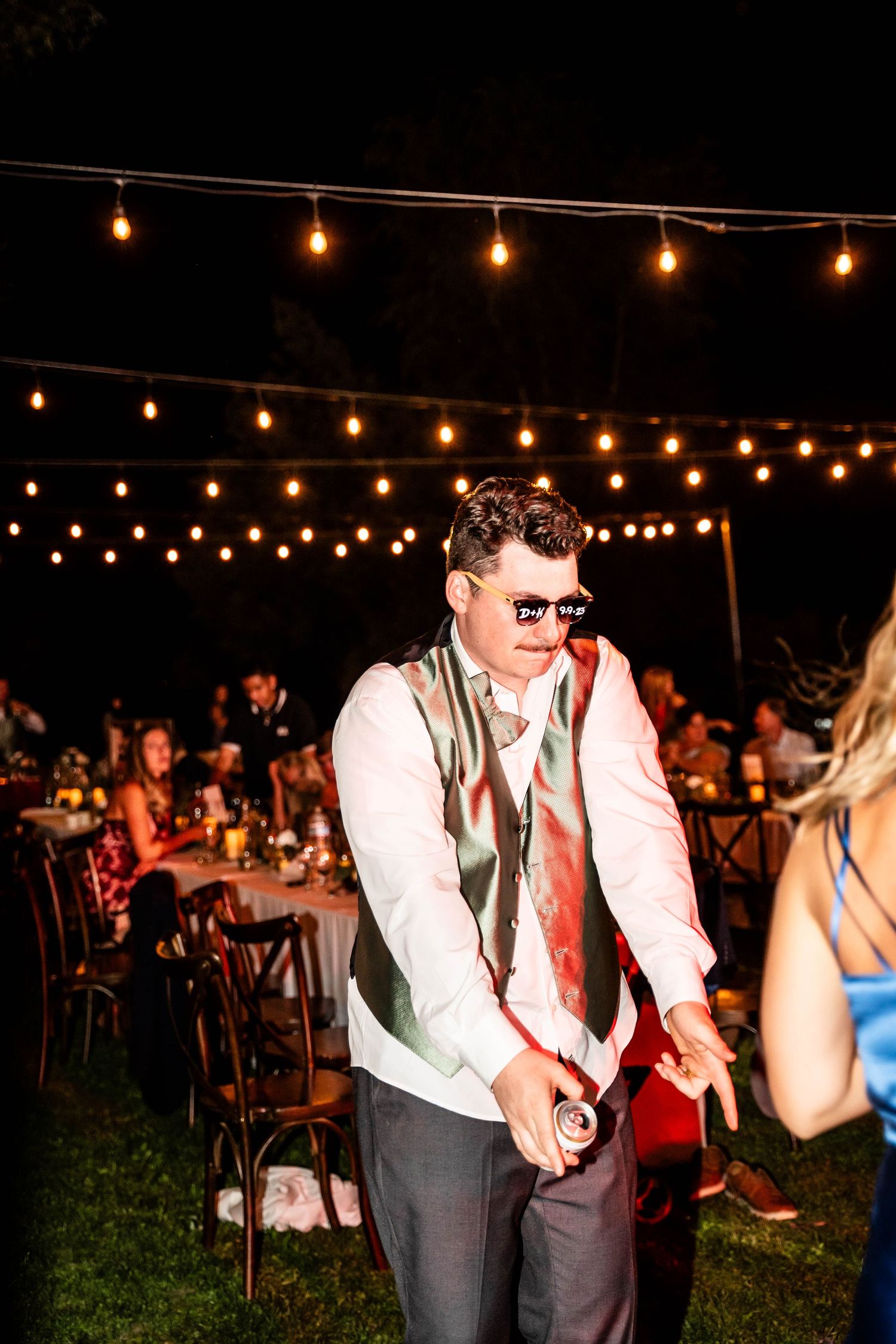 Wedding guest in sunglasses and green vest dances under string lights at night.