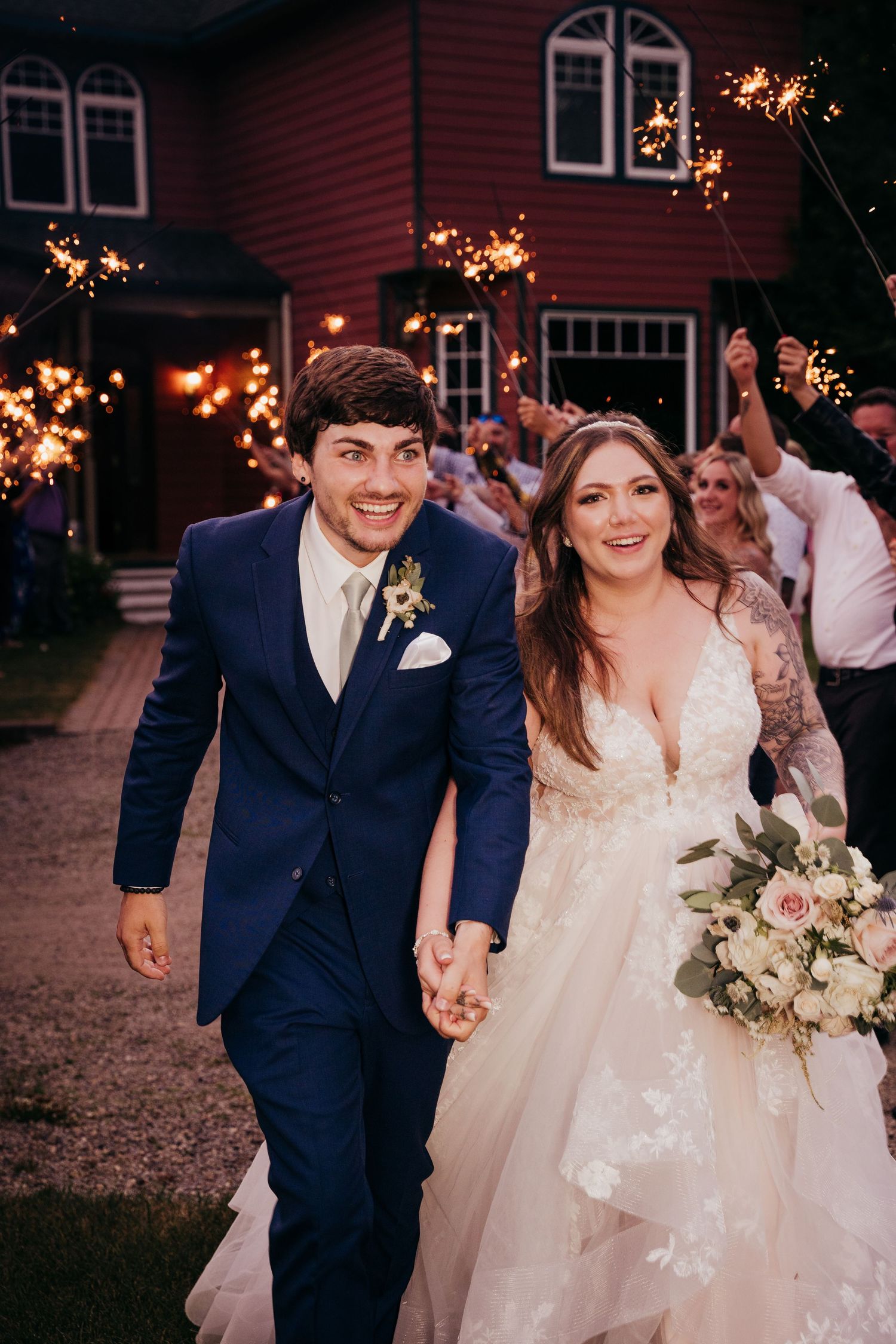 Smiling newlyweds hold hands walking through a sparkler exit at their outdoor evening wedding.