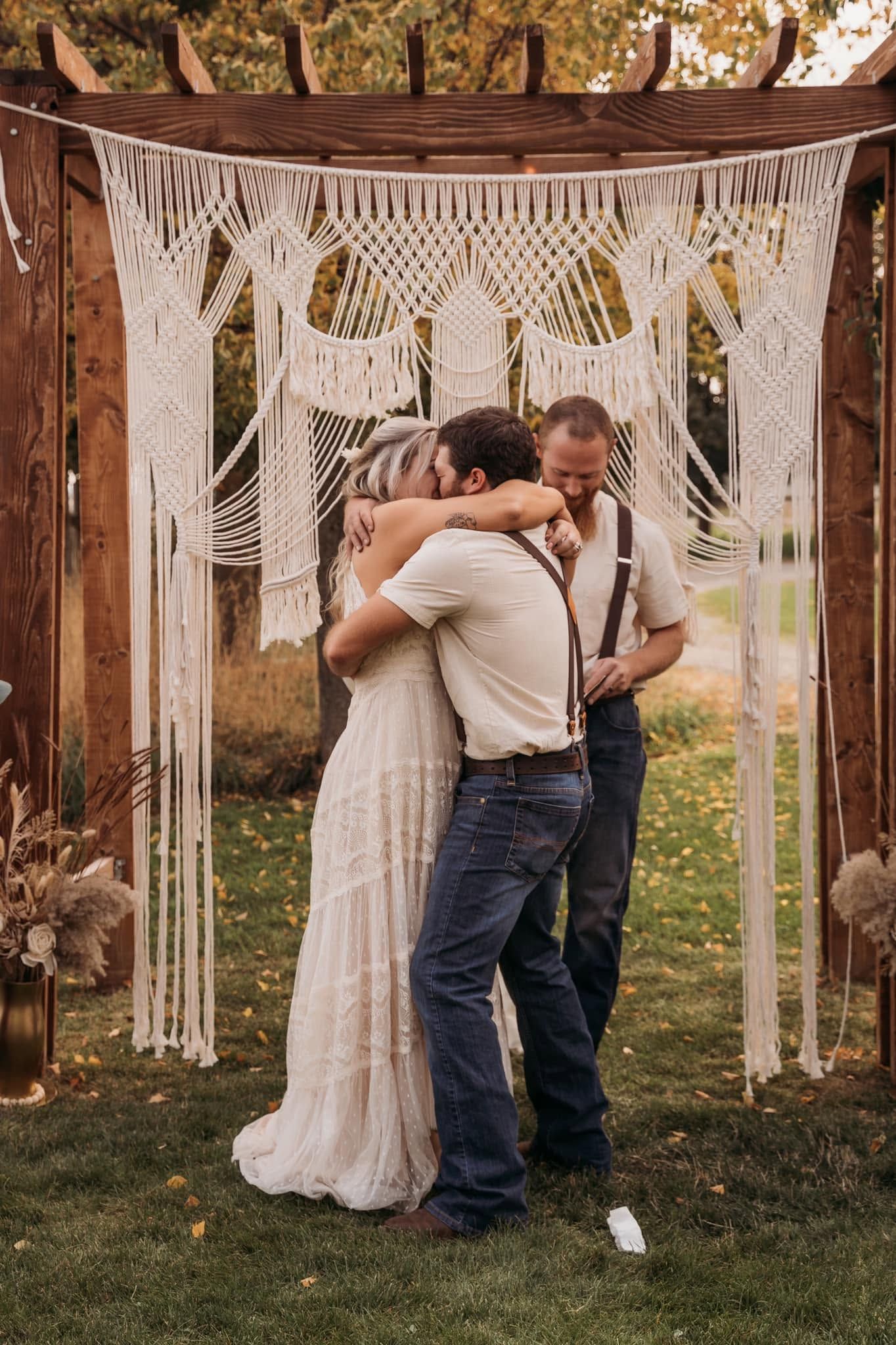 Bride and groom share an emotional three-way hug with officiant under a macrame wedding arch outdoors.