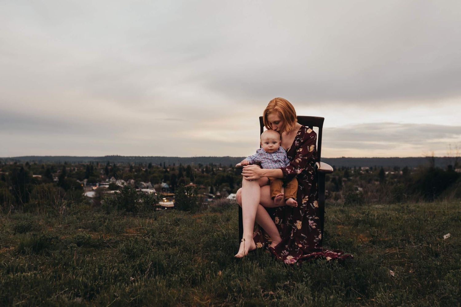 Mother sitting on chair holding baby on grassy hilltop at cloudy sunset.