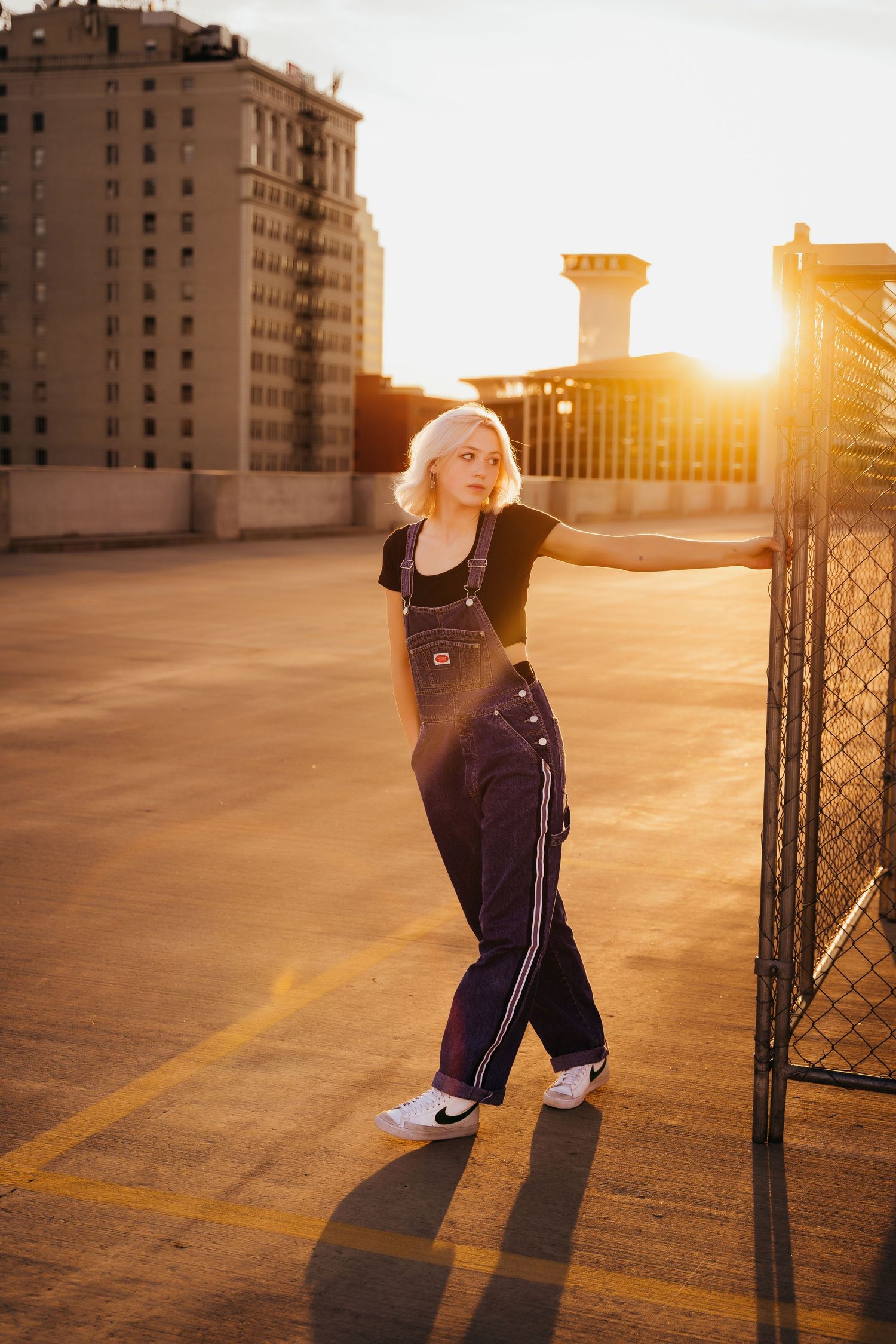 Blonde woman in denim overalls posing by chain-link fence on rooftop parking lot at sunset.