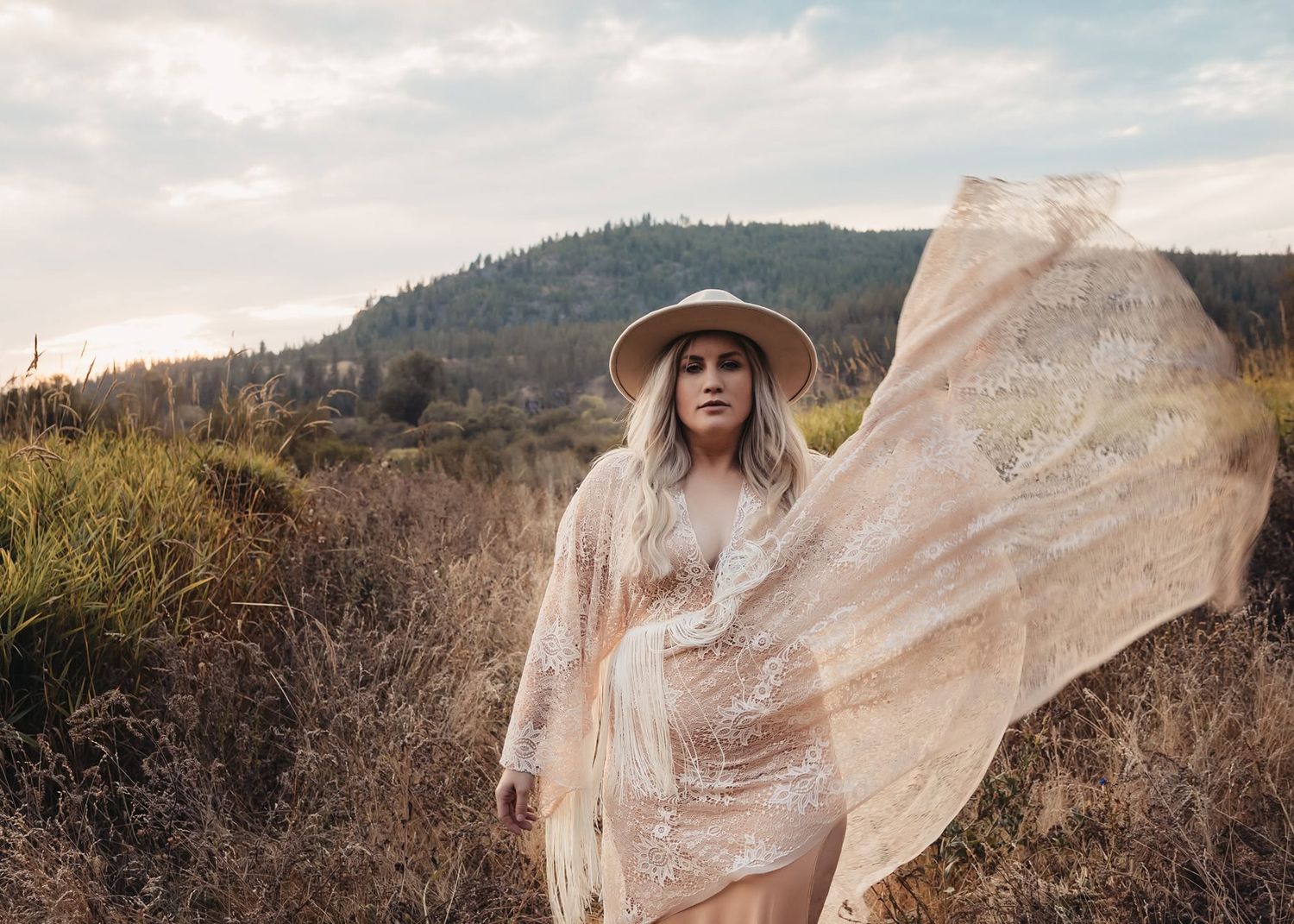 Blonde woman in lace dress and wide-brim hat stands in rustic field with fabric blowing in wind.