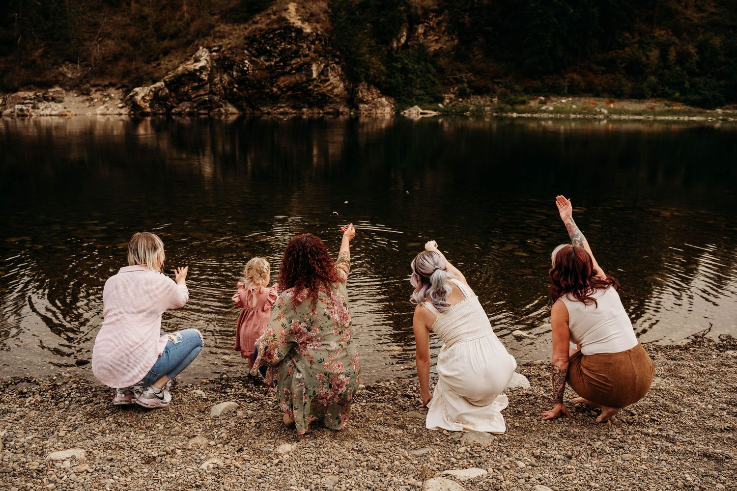 Five people crouch by a calm lake tossing stones into the water, viewed from behind with mountains in the distance.