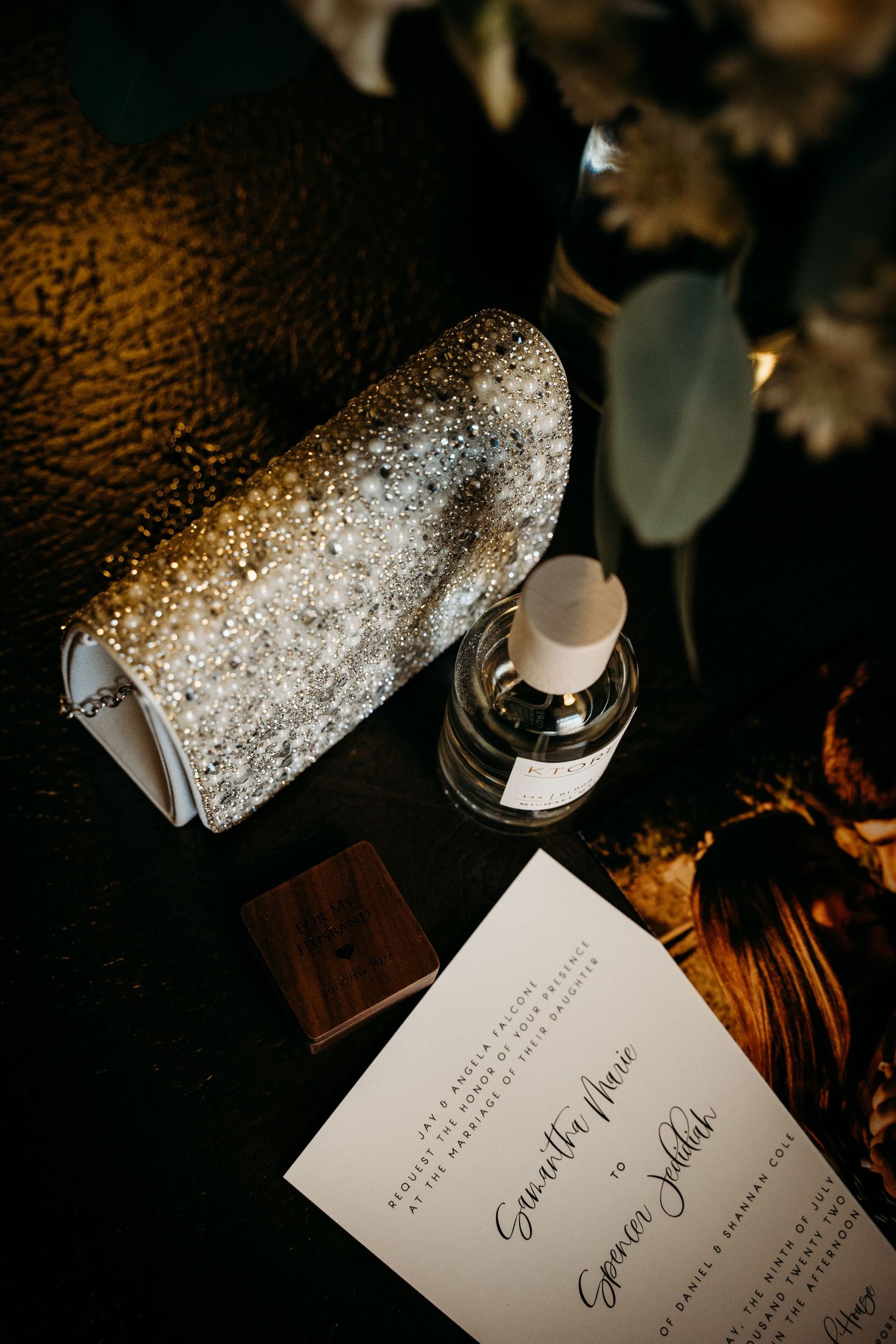 Bridal flat lay featuring glittery clutch, perfume bottle, wooden ring box, and wedding invitation.