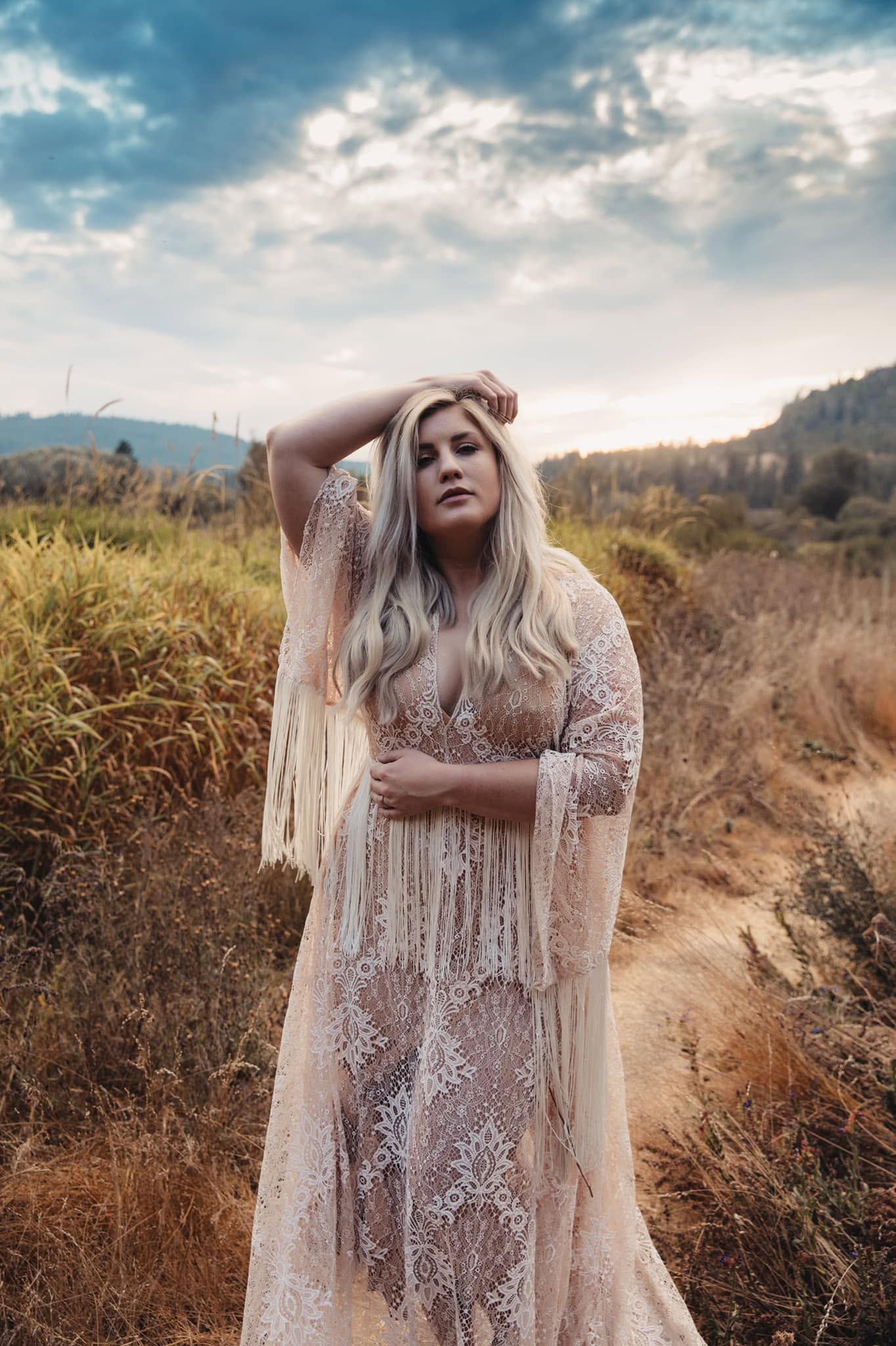 Blonde woman in a sheer lace fringe boho dress poses in a golden field under a dramatic sky.