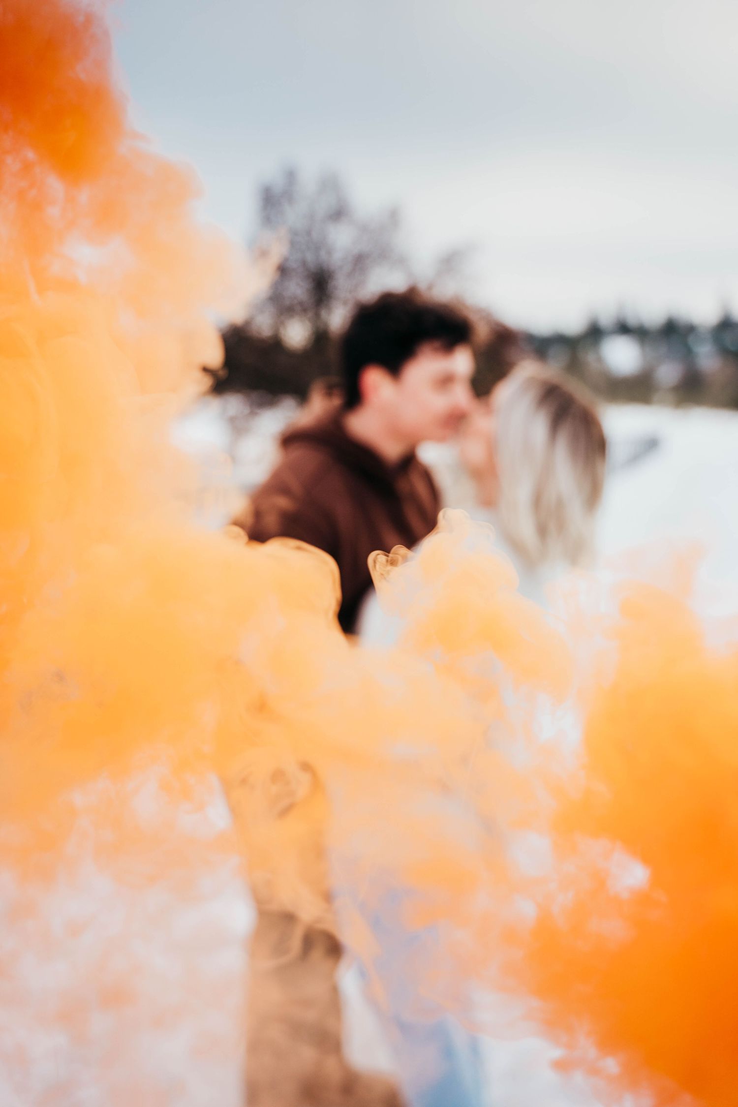 Blurred couple kissing behind vivid orange smoke bomb in a snowy outdoor scene.