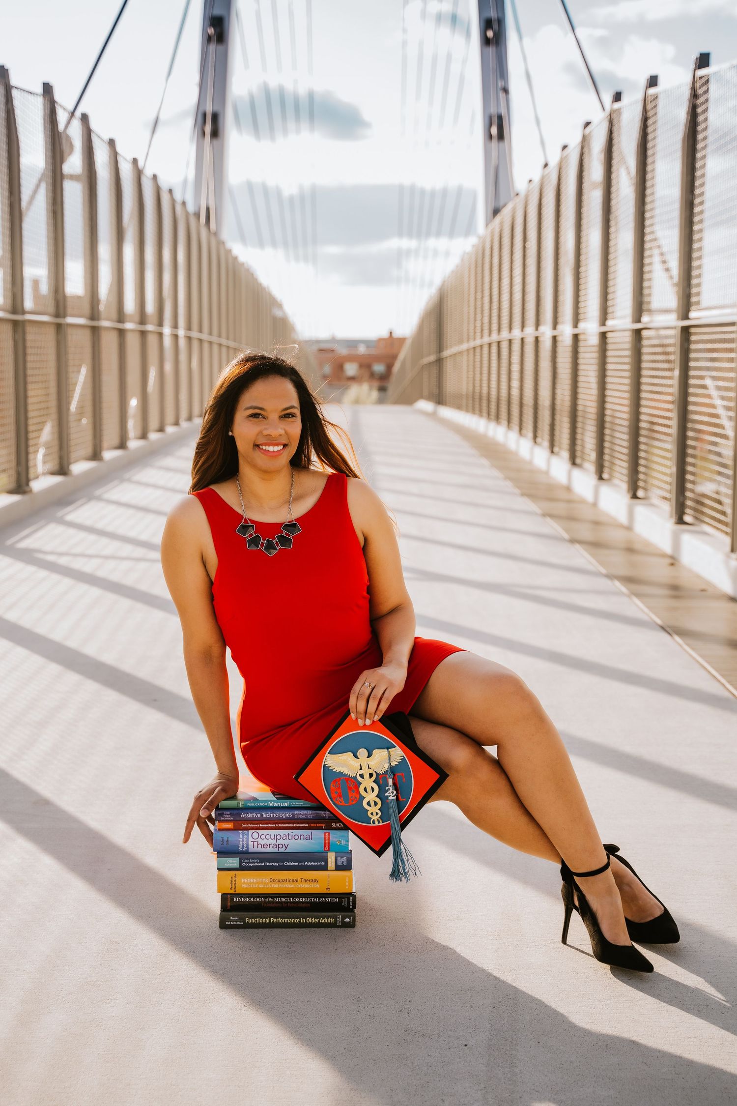 Woman in red dress sitting on bridge with graduation cap and books, celebrating medical school graduation.