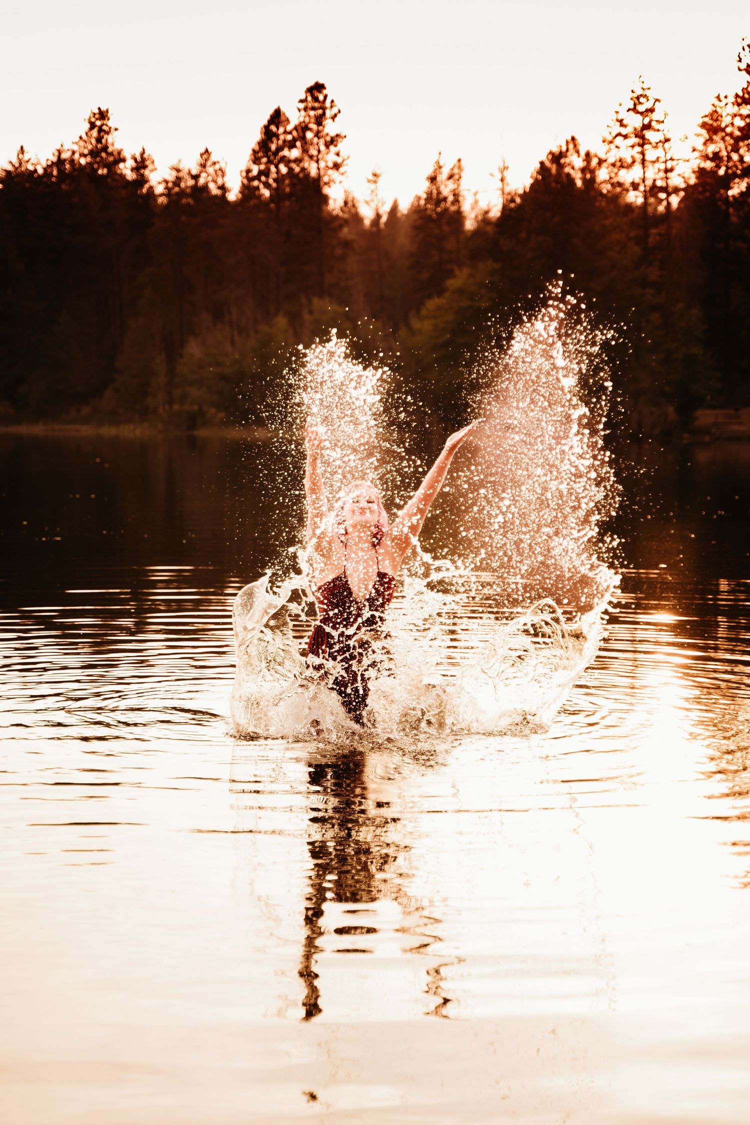 Person splashing water dramatically in a golden sunset lake surrounded by pine trees.