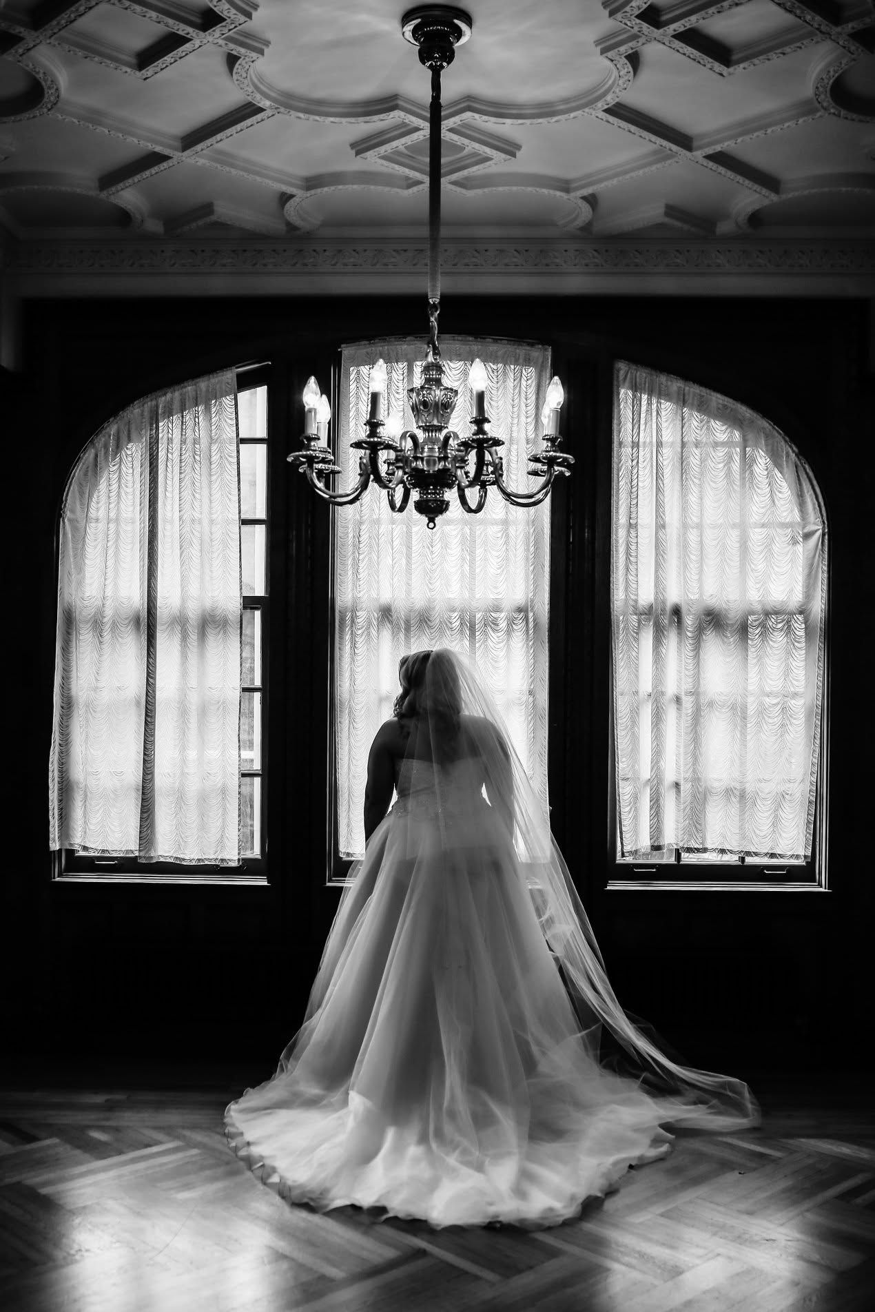 Bride in flowing gown and veil stands before arched windows in elegant ballroom.