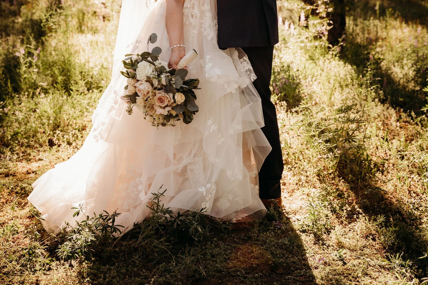 Close-up of bride in layered lace wedding gown holding bouquet of white and pink roses beside groom in forest.