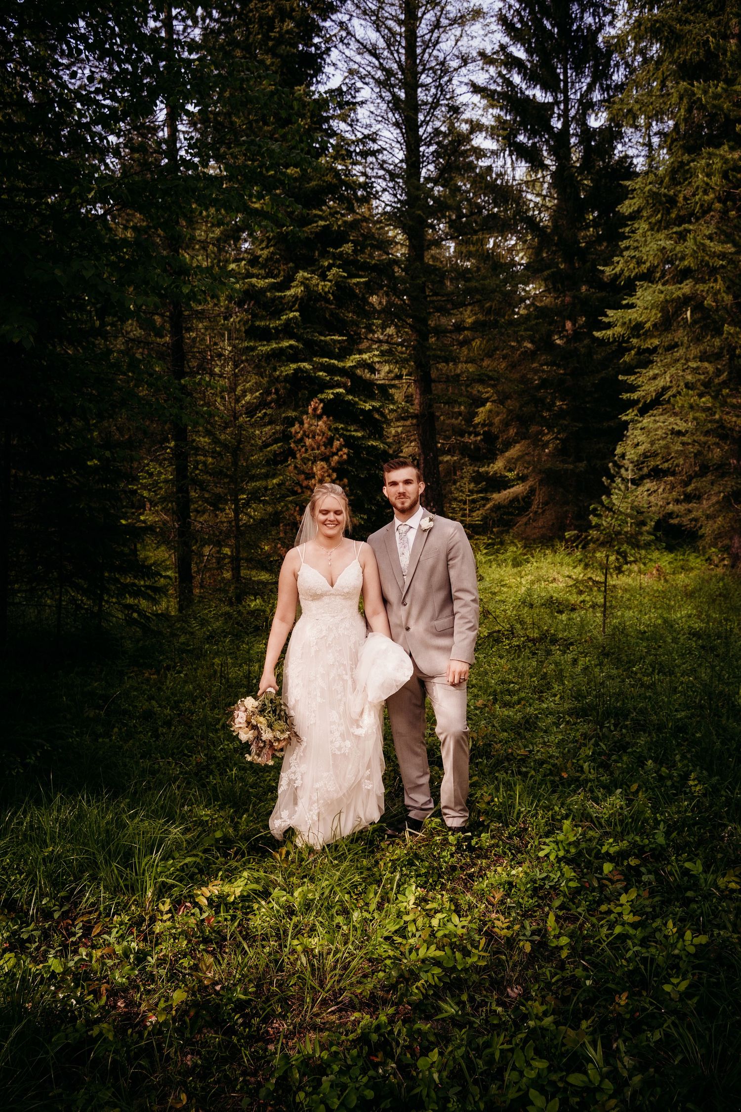 Bride and groom posing together in a lush green forest, bride in lace gown with veil.
