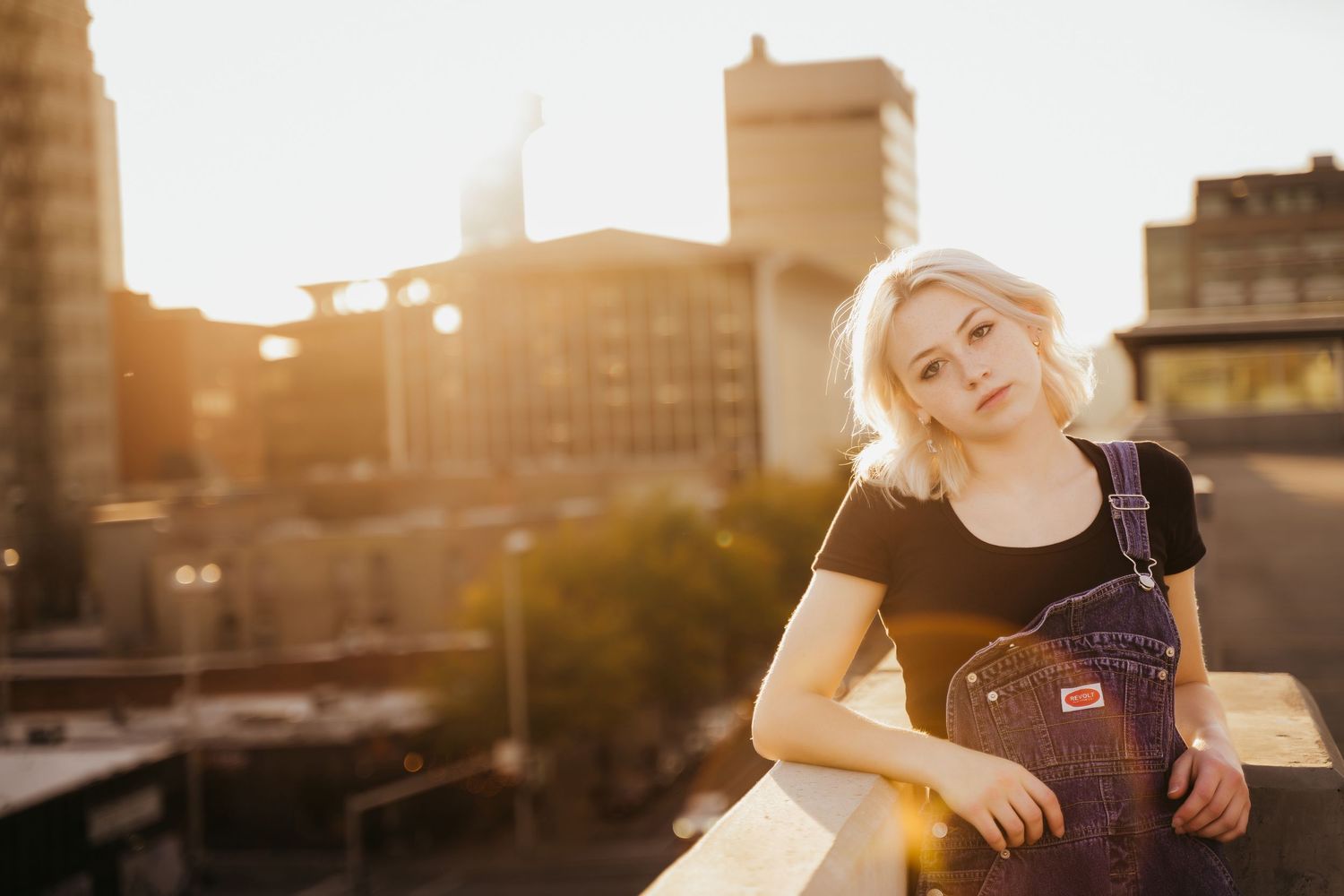 Young blonde woman in denim overalls leans on a rooftop railing during a glowing urban sunset.