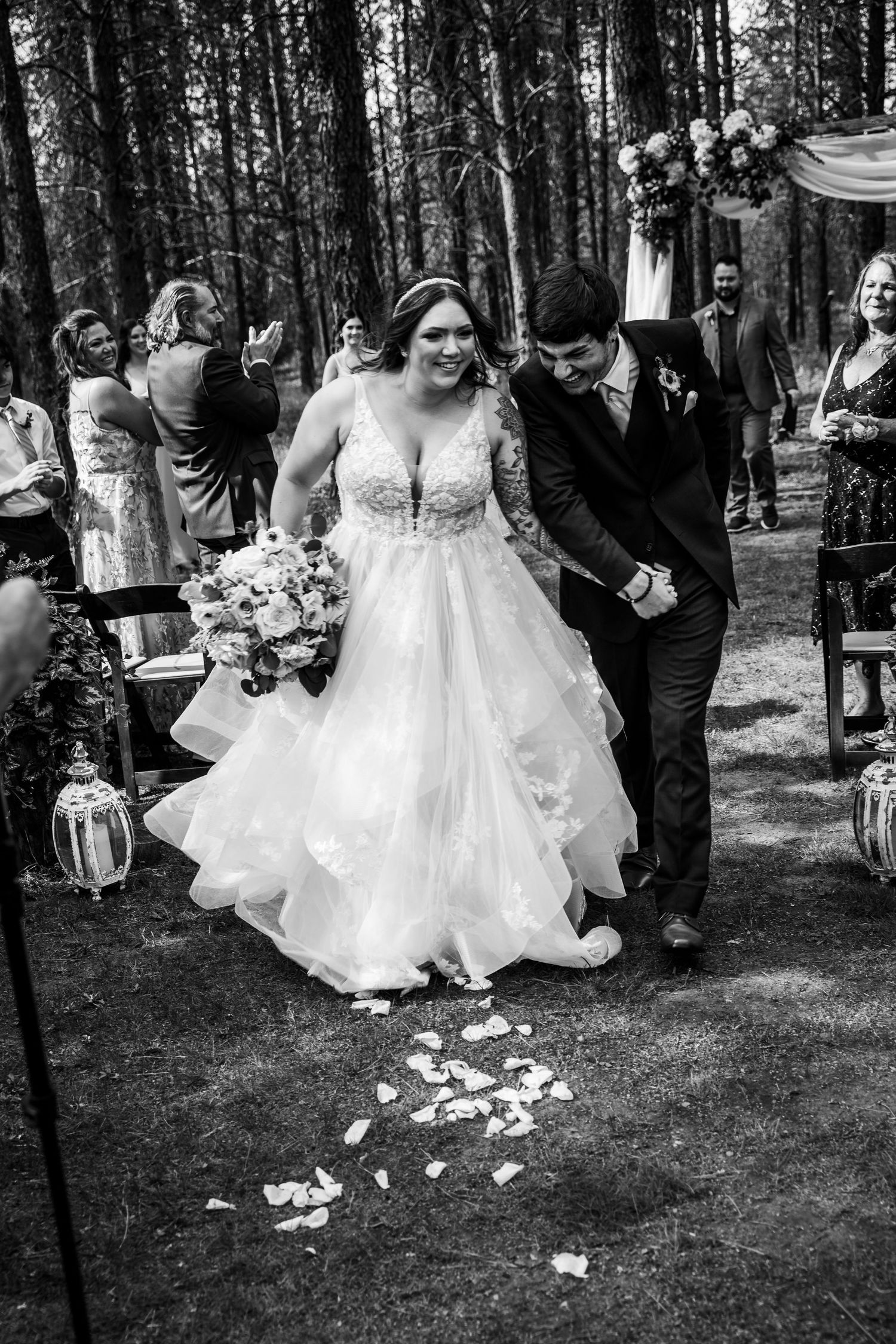 Black and white photo of newlyweds walking down petal-lined aisle after forest ceremony.