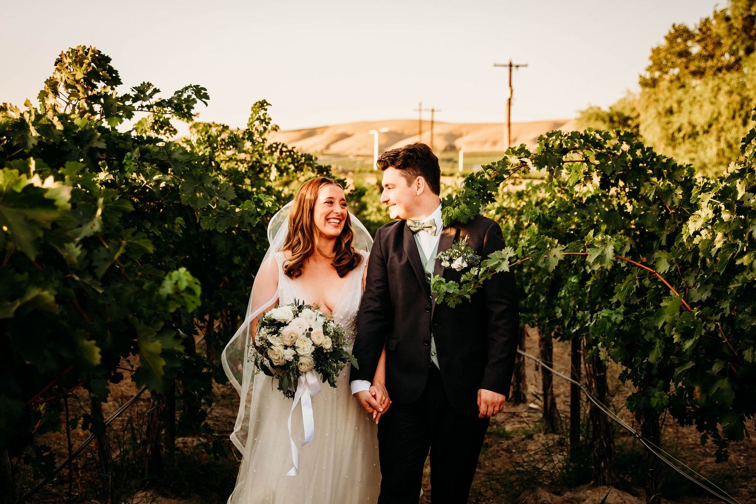 Smiling newlyweds walk hand in hand through a vineyard holding a white bouquet.