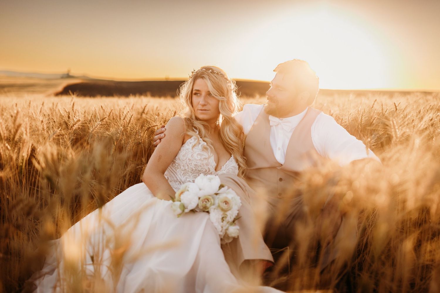 Bride and groom sit together in golden wheat field during sunset wedding portraits.