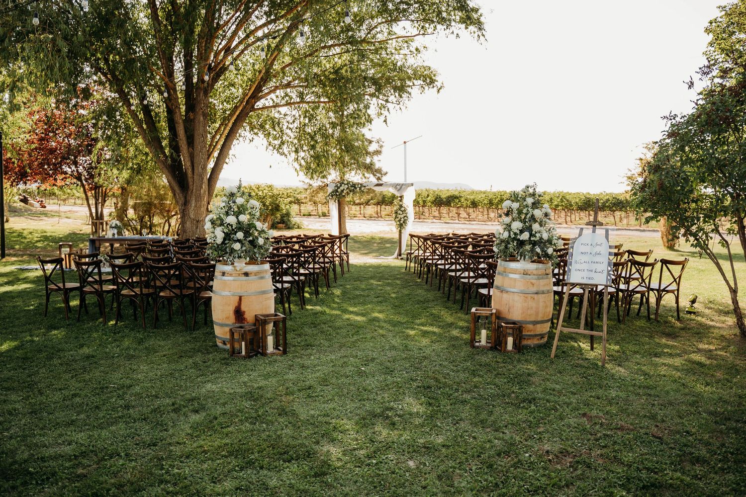 Outdoor wedding ceremony setup with wooden barrel aisle markers, floral arch, and rows of dark wooden chairs.