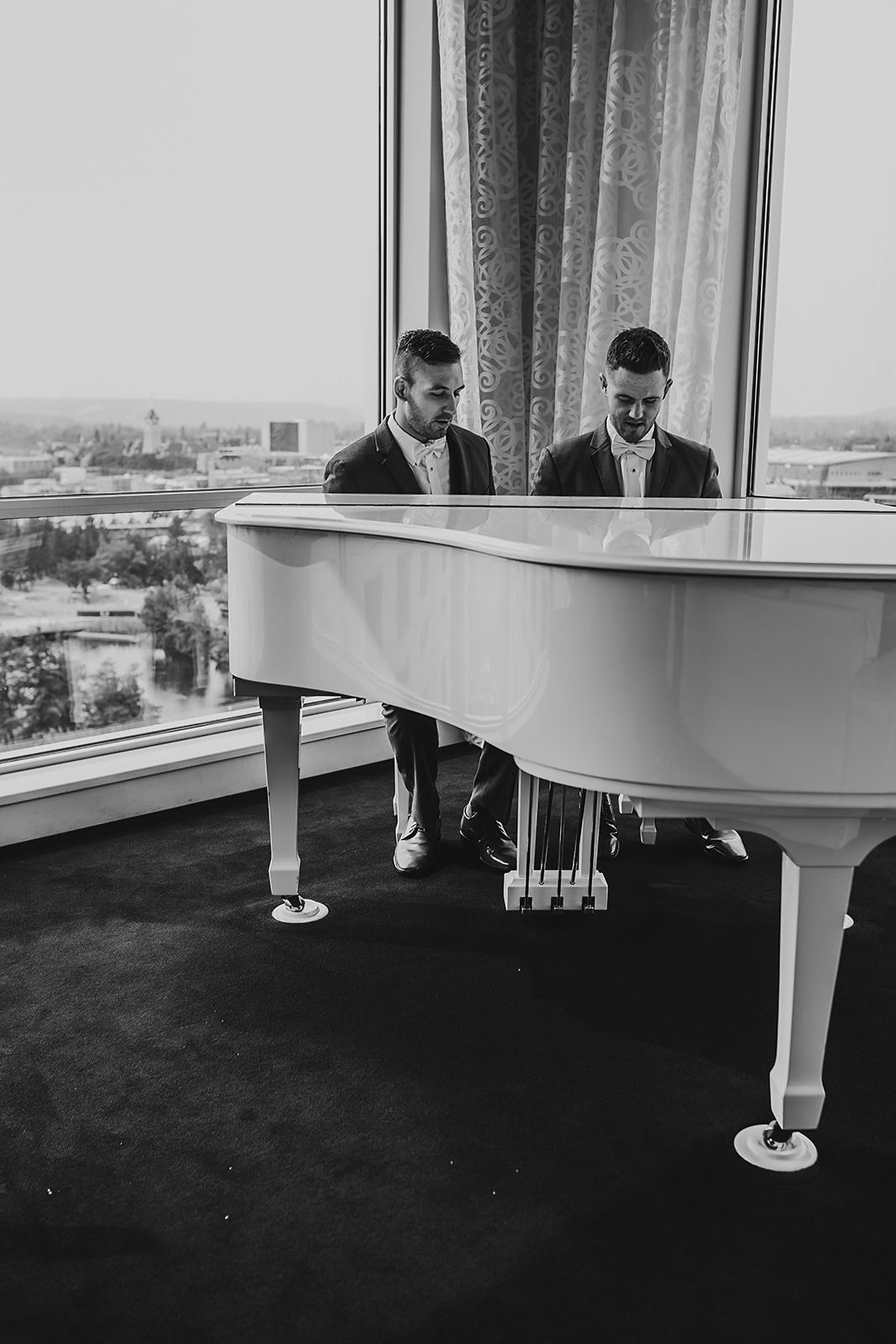 Two grooms in black suits sit together at a white grand piano in a hotel suite.