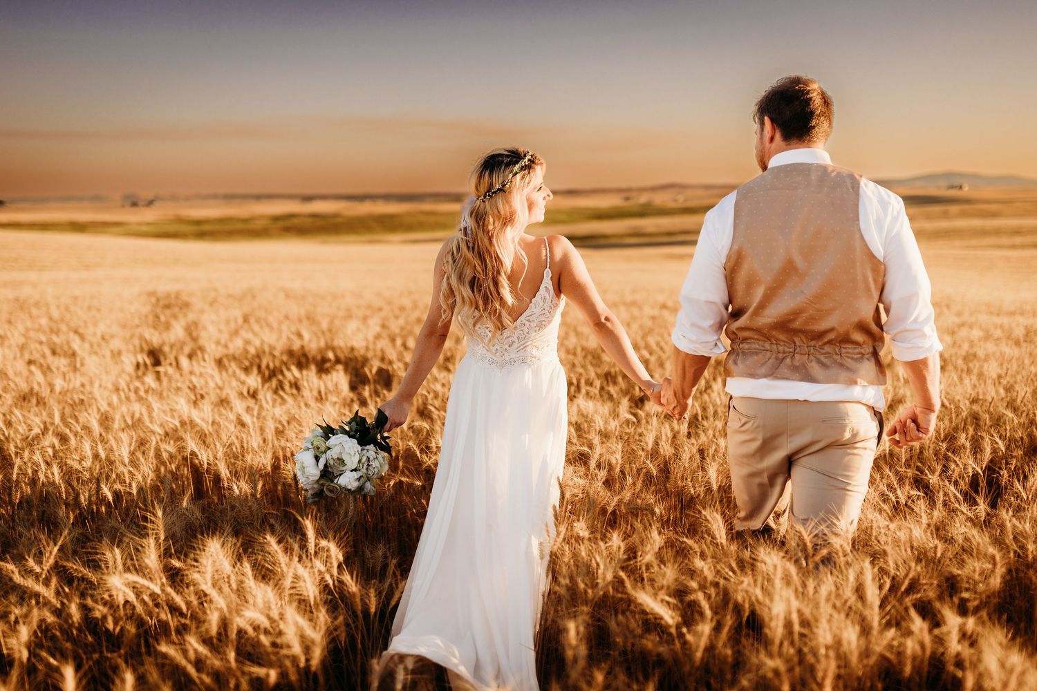 Bride and groom walk hand in hand through golden wheat field at sunset.