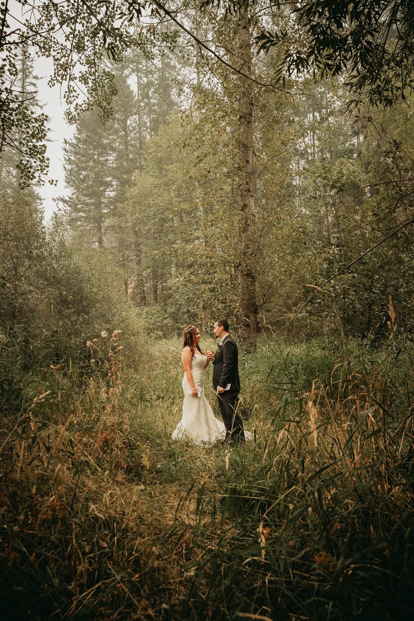 Bride in white gown and groom in dark suit face each other on a misty forest path surrounded by wildflowers.