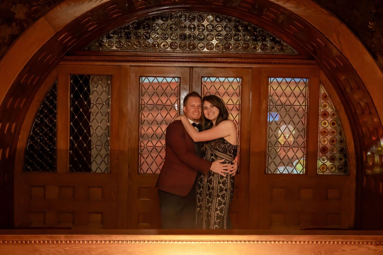 Couple embraces under an ornate wooden arch with stained glass windows in a warmly lit venue.