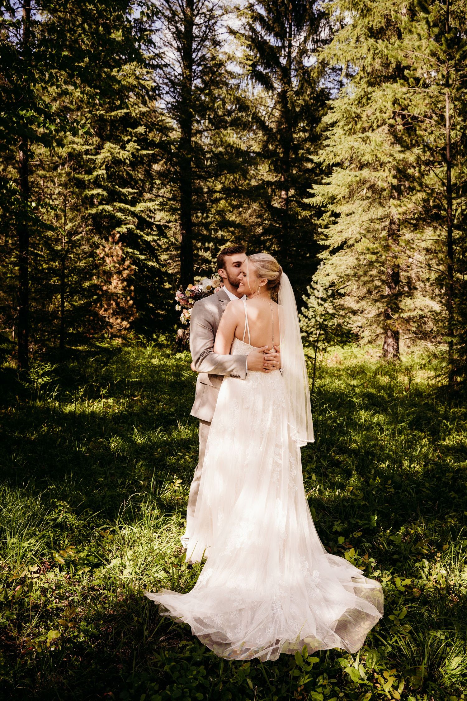 Bride and groom kissing in a lush green forest, bride in elegant lace gown with long veil.