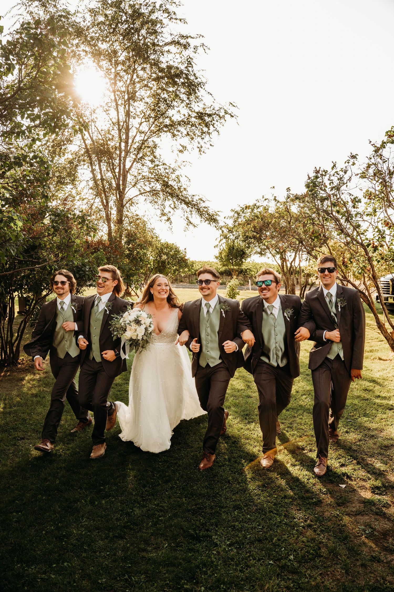 Bride poses with groomsmen in dark suits and sage vests wearing sunglasses in sunny outdoor setting.