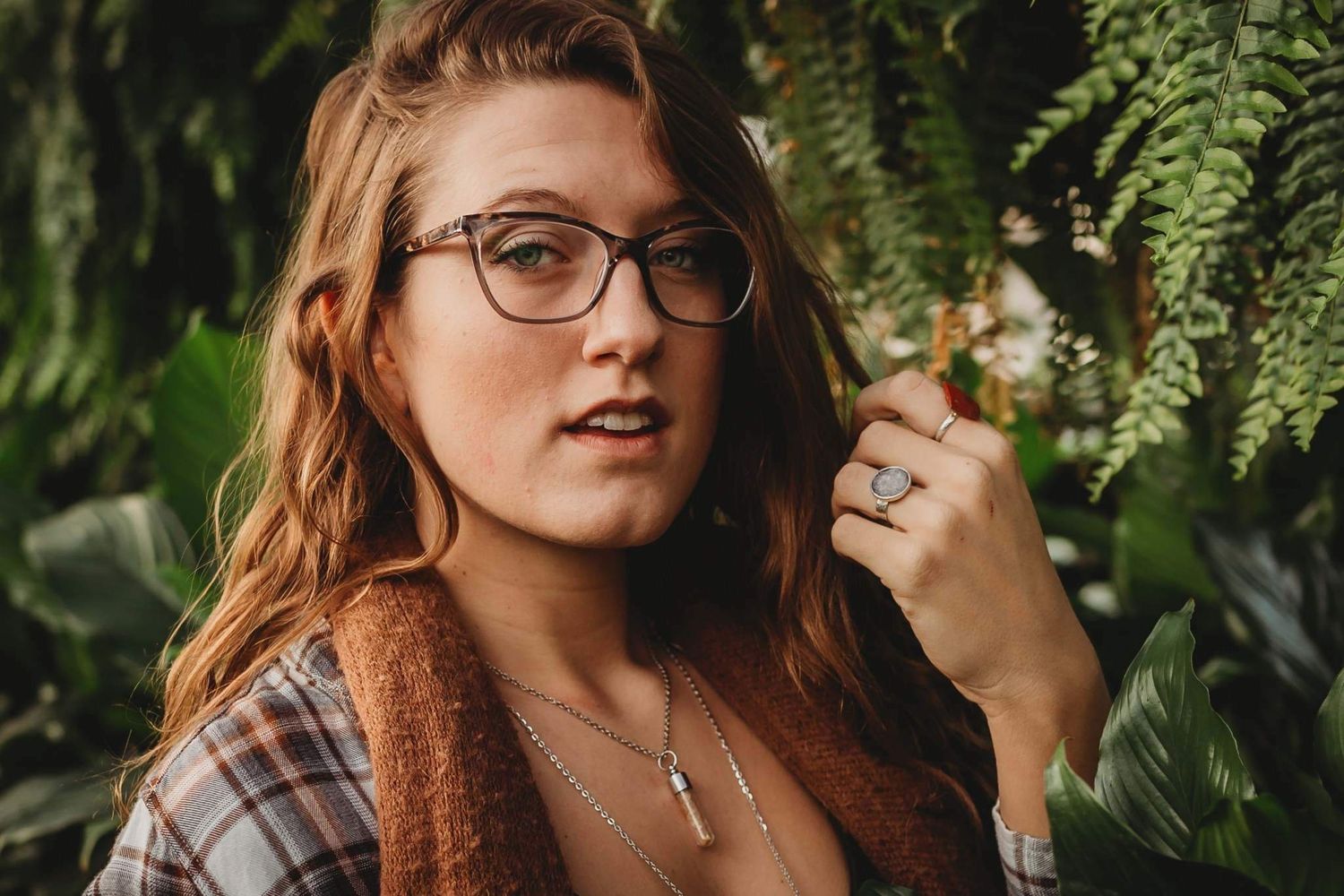 Young woman with glasses and auburn hair poses among lush green ferns wearing layered necklaces.