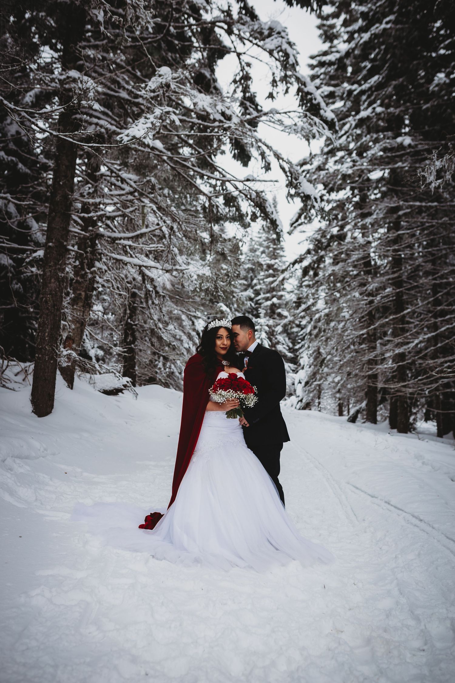 Bride in red cape and groom stand in snow-covered pine forest wedding photo.