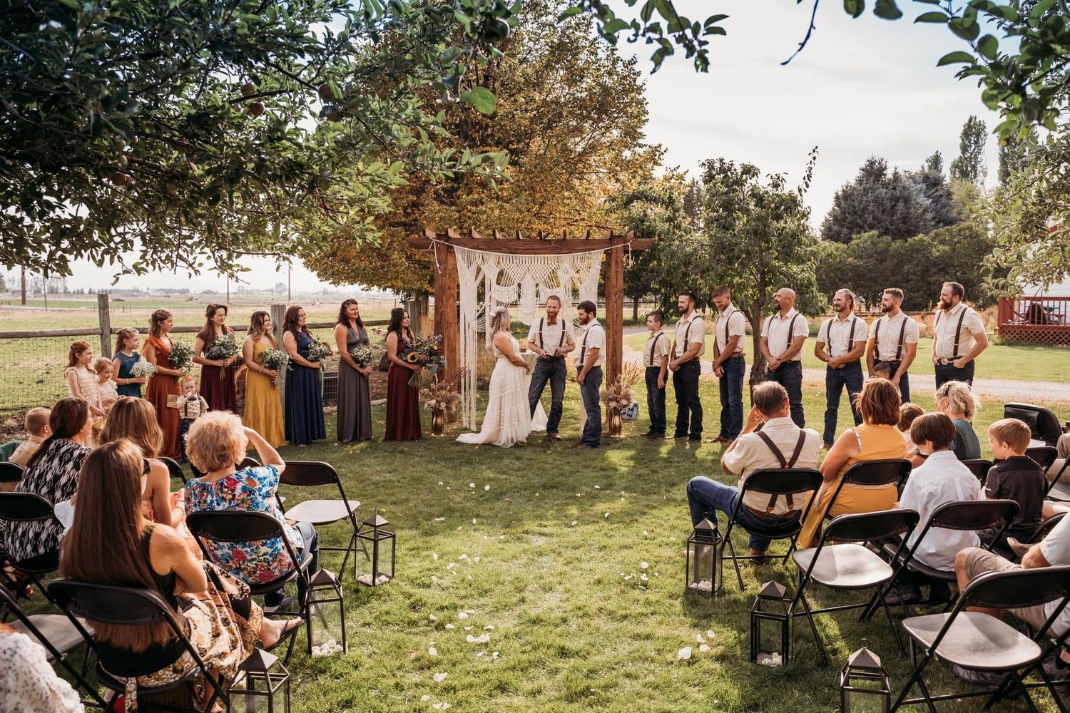 Outdoor boho wedding ceremony under wooden arch with macrame, bridal party on green lawn.