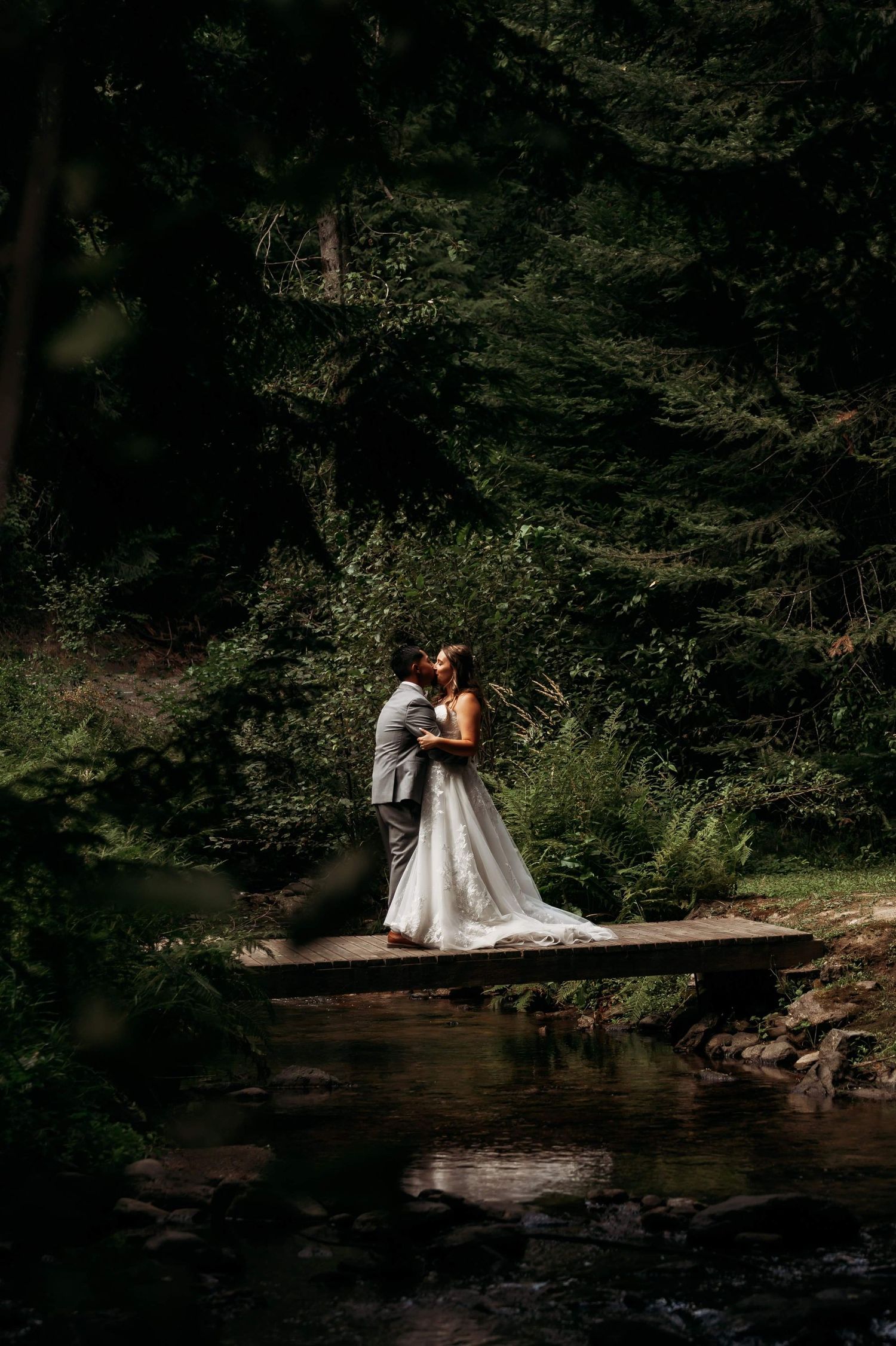 Bride and groom sharing a kiss on a wooden bridge over a stream in a lush green forest.