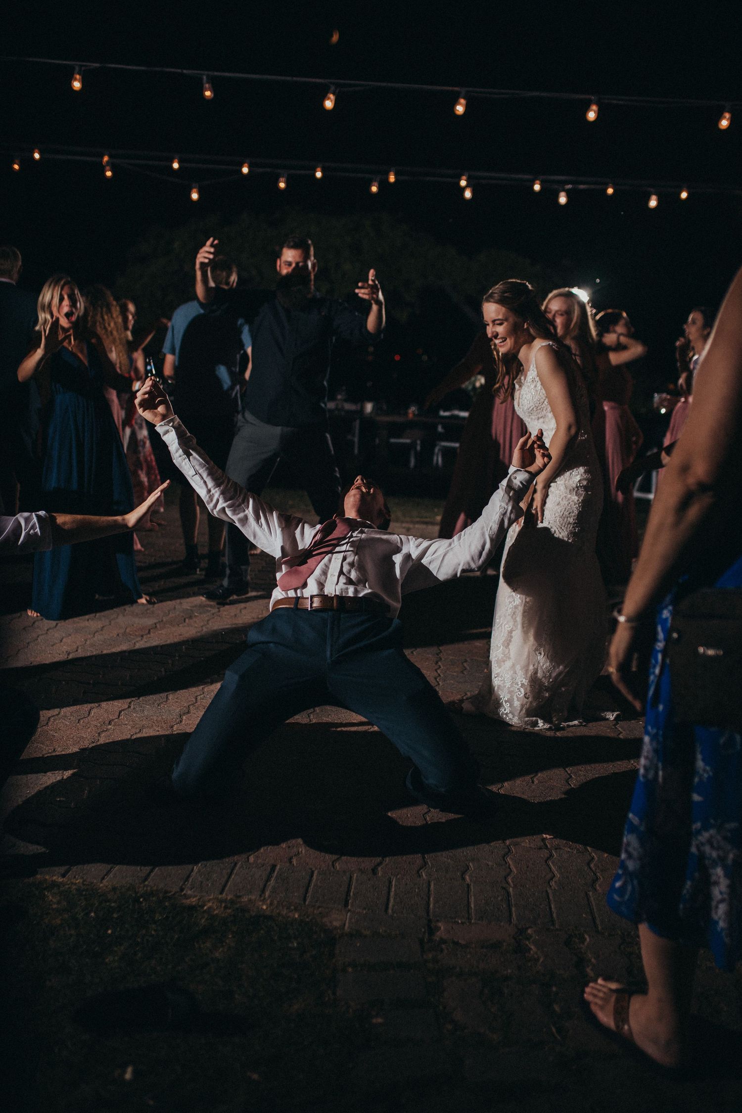 Groom slides on his knees on the dance floor as bride laughs during lively wedding reception.
